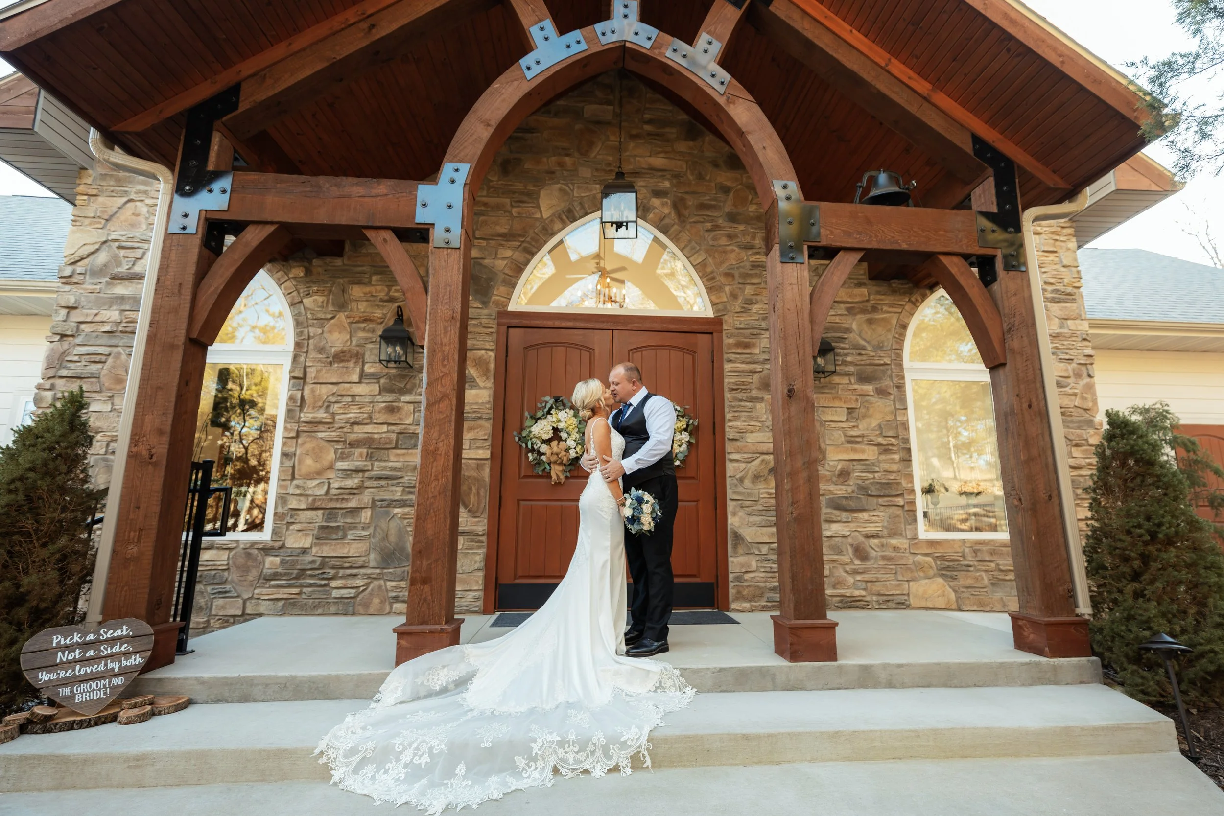 A bride and groom are standing close together in front of a house with a stone exterior and wooden archway. The bride is wearing a white wedding gown with a long train, and the groom is in a dark vest and white shirt. They are holding each other and 