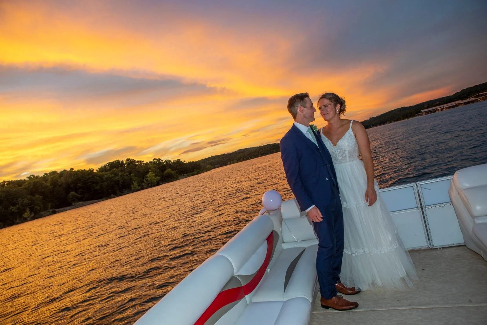 A couple in wedding attire standing on a boat during sunset, with a river and silhouetted trees in the background.