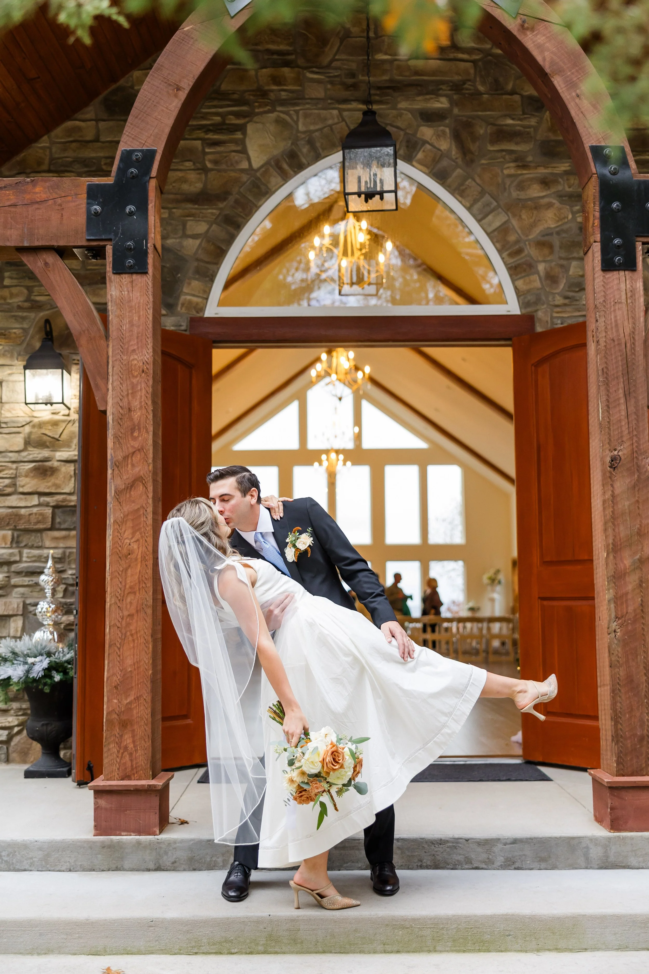 A newlywed couple sharing a romantic moment outside a church. The groom is dipping the bride as they kiss, with the bride holding a bouquet of flowers. The couple is standing in the archway of the church, which has a stone facade and wooden doors.