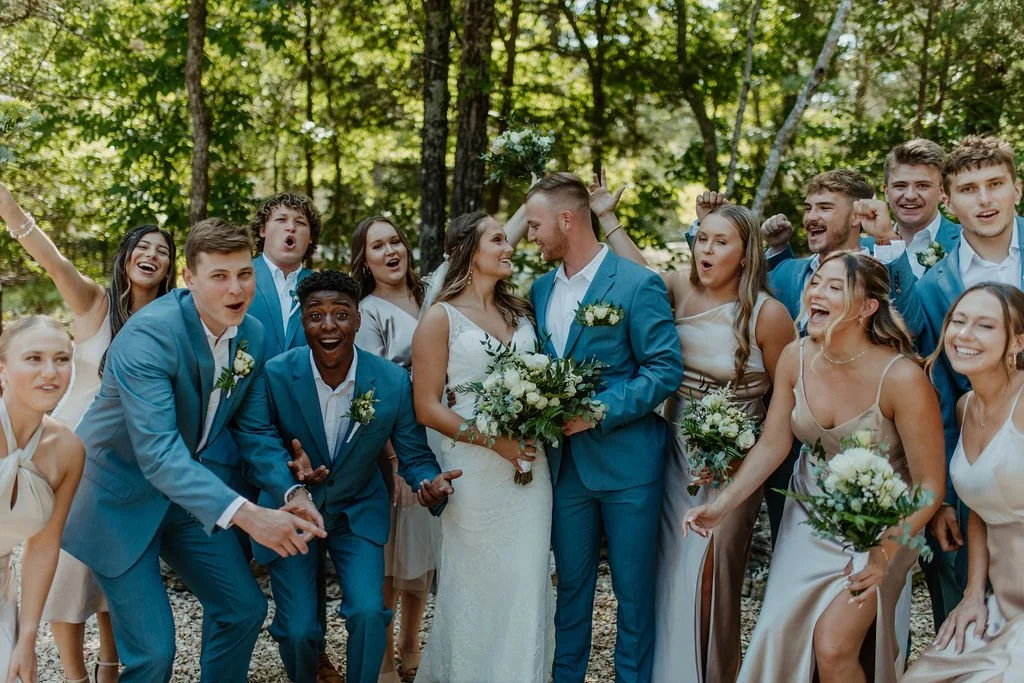 A wedding celebration outdoors with a bride and groom surrounded by smiling wedding guests in a wooded area.
