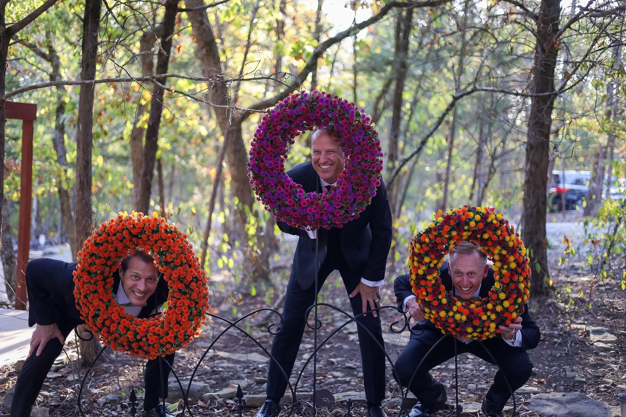 Four men in suits participating in a celebratory event outdoors during autumn, with each man holding a vibrant flower wreath around their faces, smiling, among trees with sunlight filtering through the leaves.