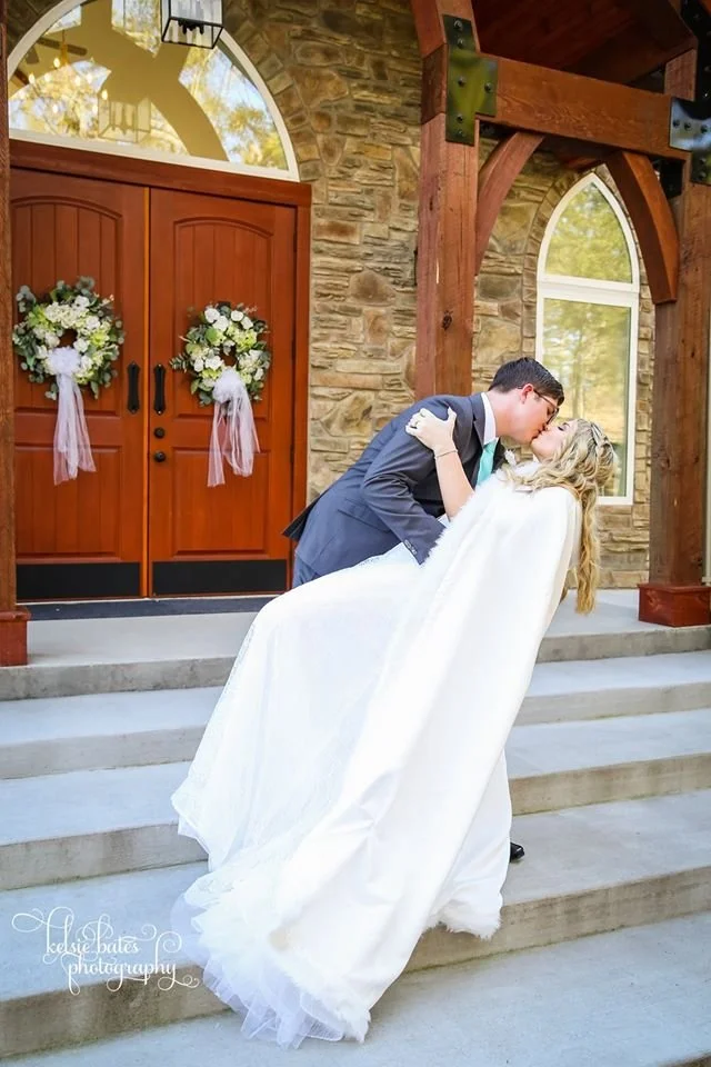 A newlywed couple sharing a kiss on the steps of a stone building decorated with floral wreaths and pink ribbons, during a wedding ceremony or celebration.