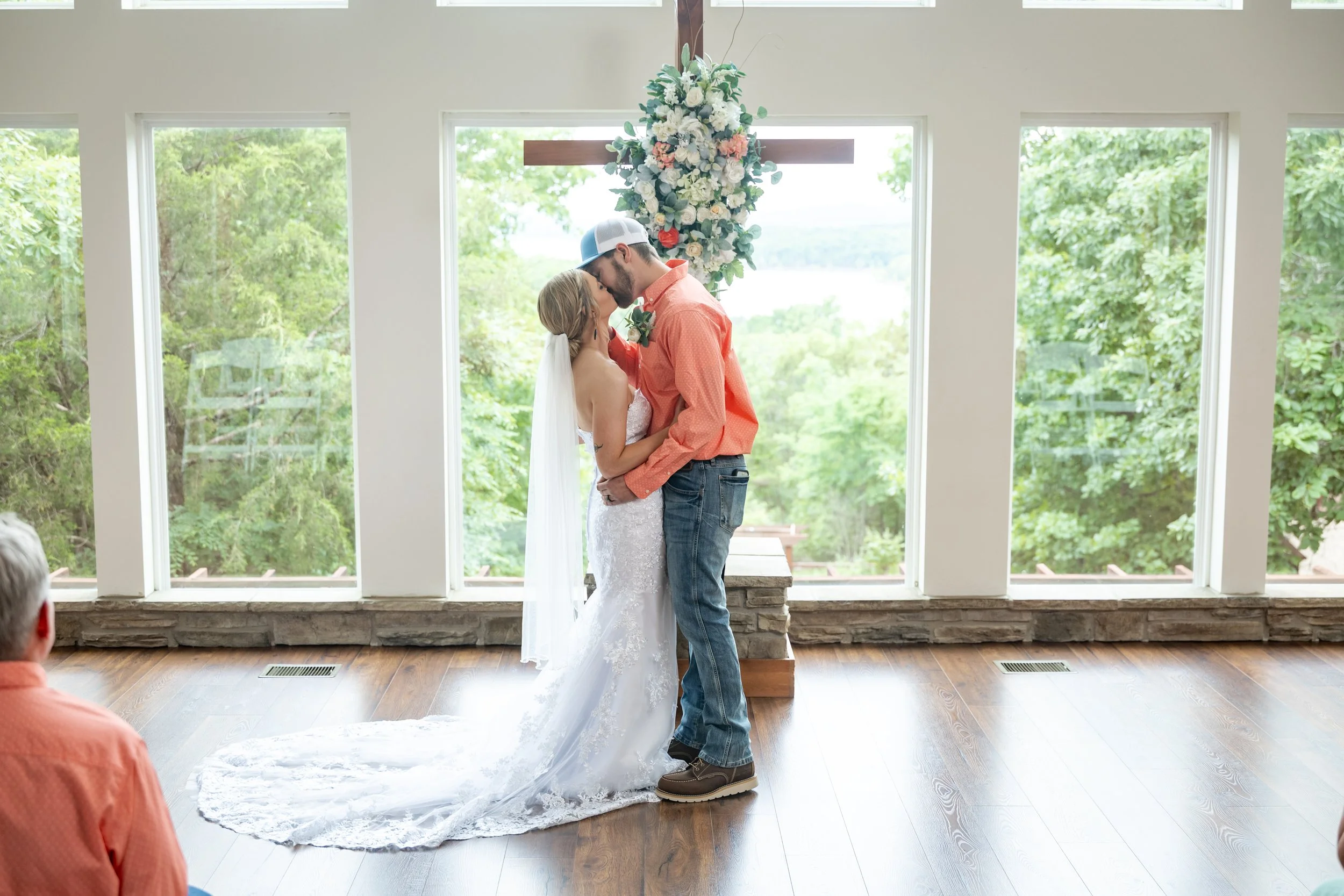 A bride and groom are sharing a kiss during their wedding ceremony inside a venue with large windows showing green trees outside, standing beneath a cross decorated with white and pink flowers.
