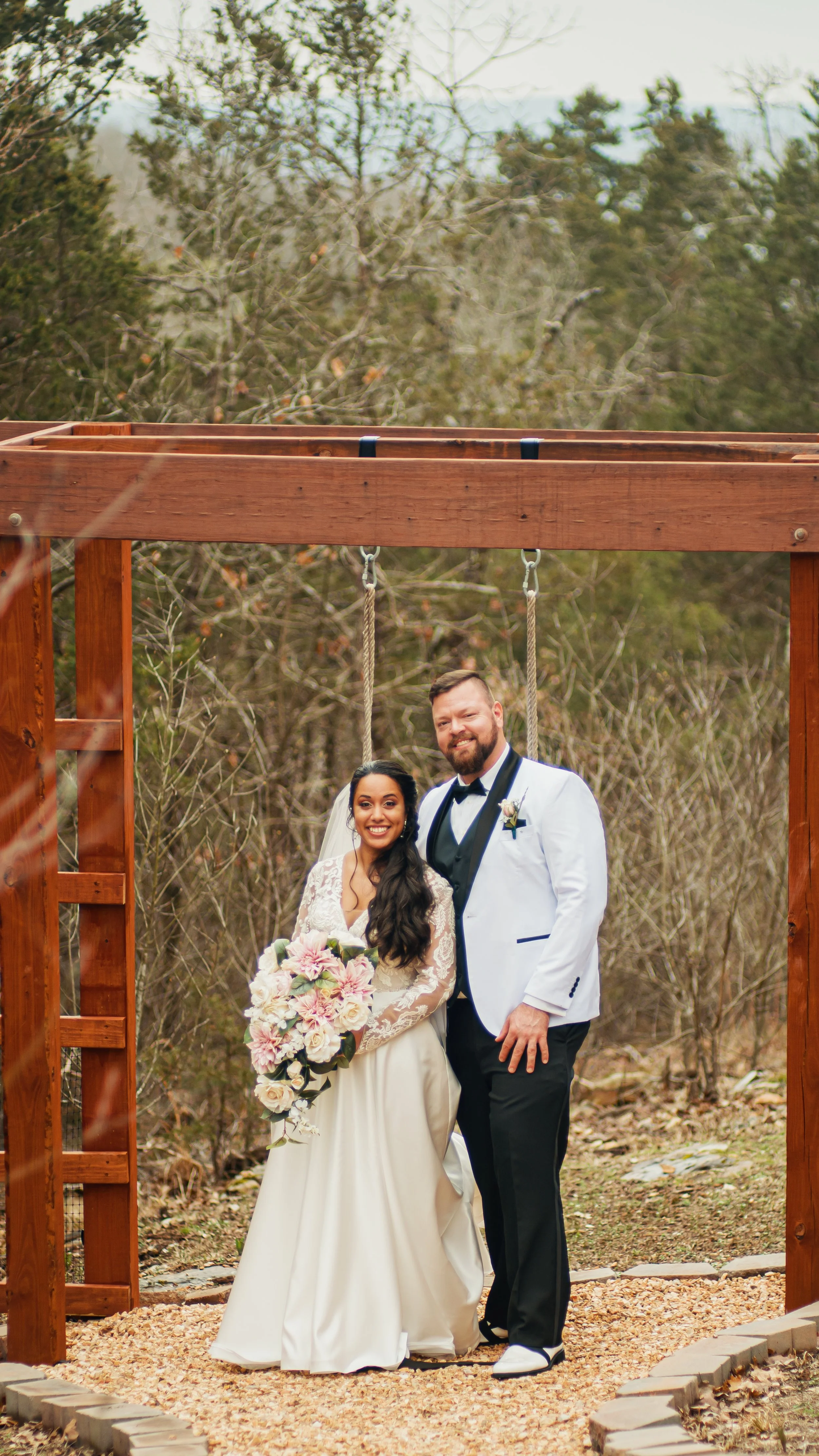A bride and groom standing under a wooden arch on their wedding day outdoors; the bride is holding a large bouquet of flowers, and both are smiling.
