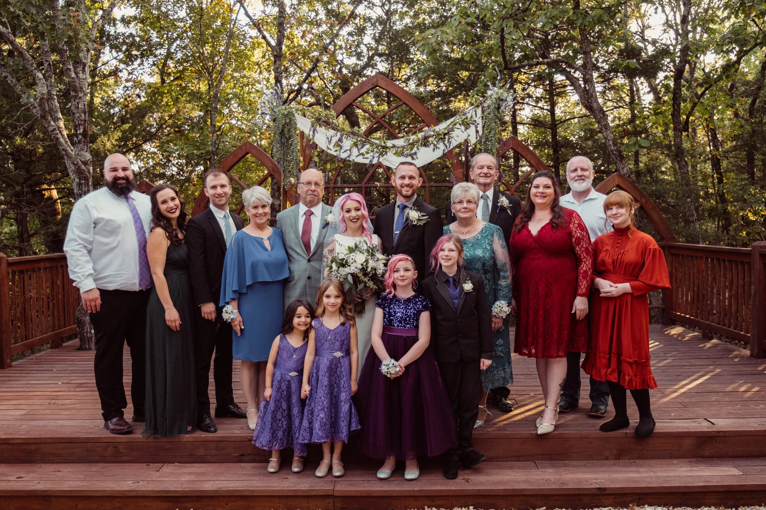 Group of people at a wedding, including a bride with pink hair, groom, children, and family members, standing on a wooden deck in a forested outdoor setting with decorative greenery and drapery overhead.