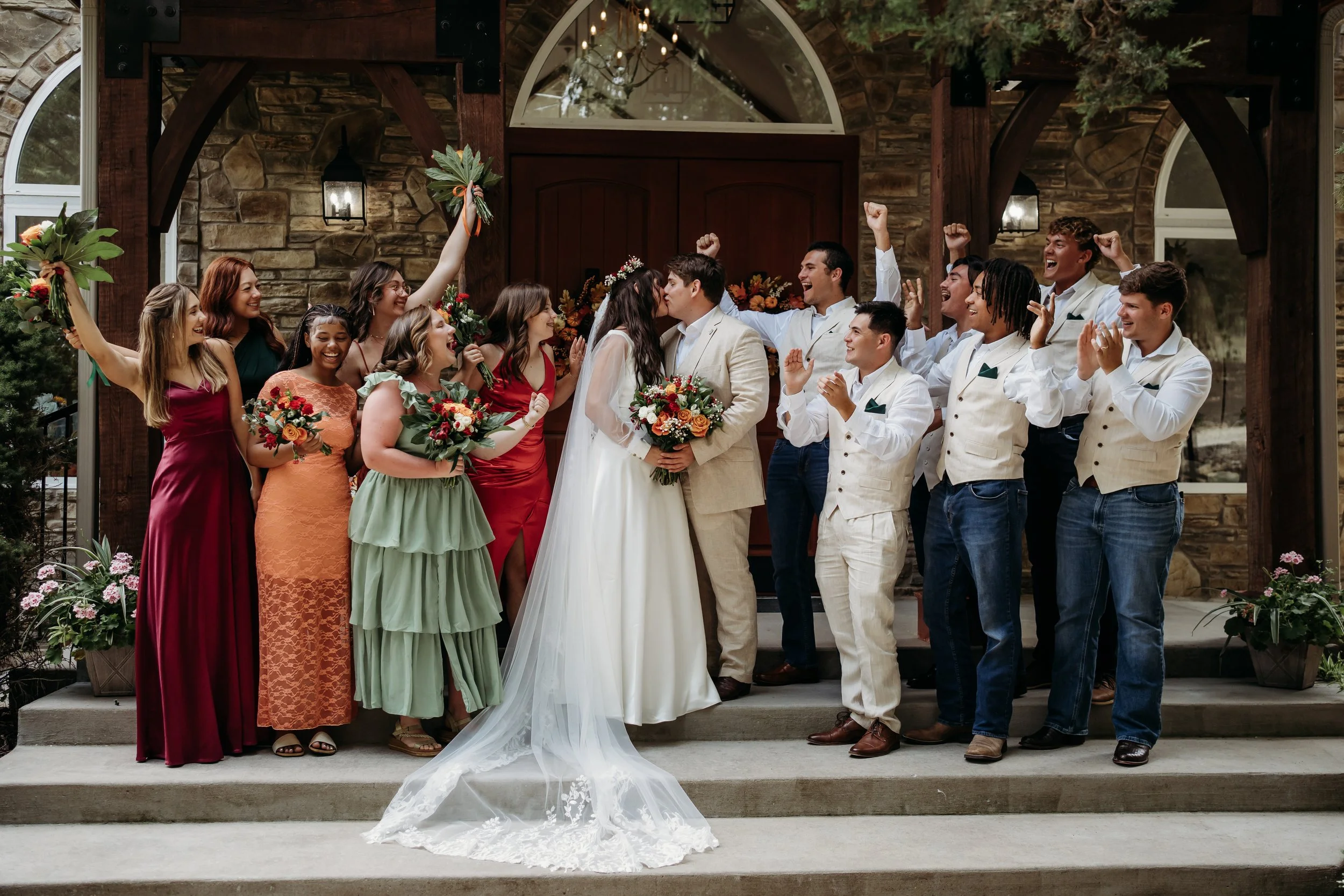 Celebrating a wedding on steps outside a stone building with a wooden arch. The bride and groom are kissing, surrounded by bridesmaids and groomsmen. Some people are cheering and raising their fists. The scene is joyful and festive.