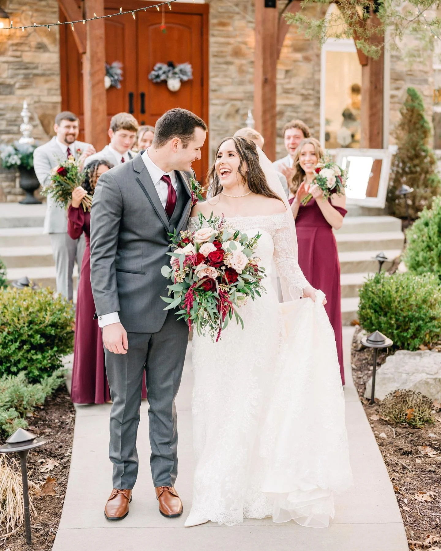 A bride and groom are smiling at each other while standing on a wedding aisle. The bride is holding a large bouquet of flowers, and the groom is dressed in a gray suit. In the background, wedding guests are clapping and smiling, standing in front of 