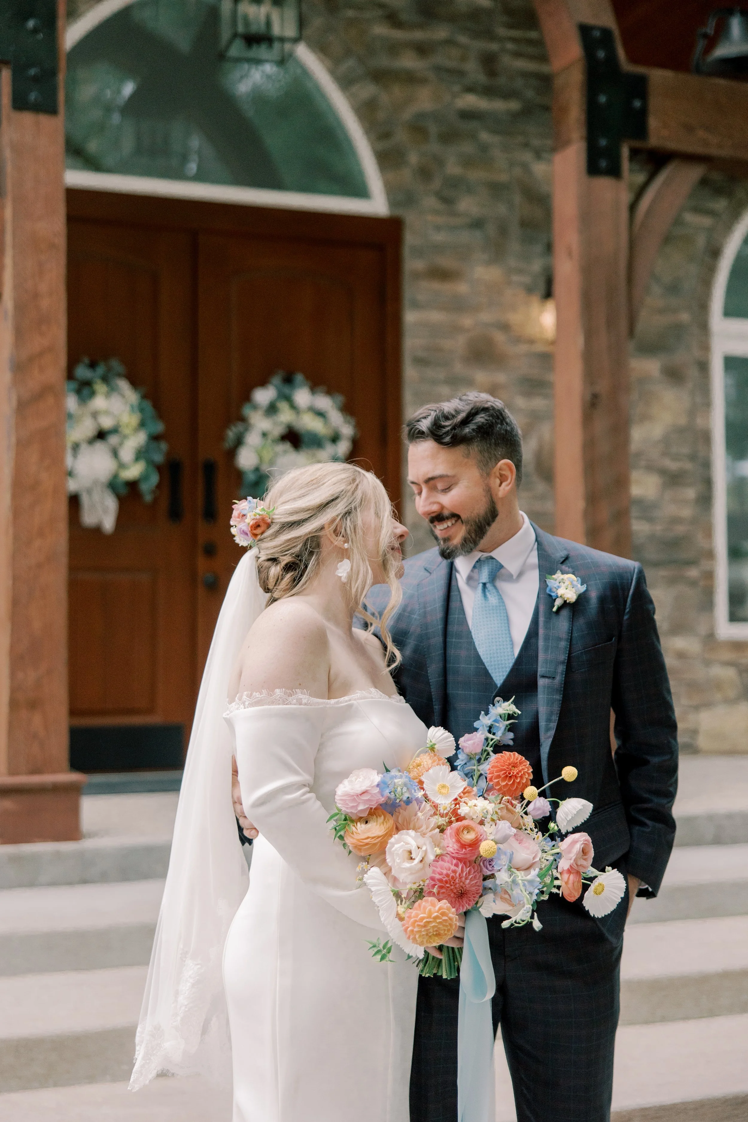 A bride and groom smiling at each other during their wedding ceremony inside a church with stone walls and large arched windows. The bride is holding a colorful bouquet and wearing a white dress with lace detail, while the groom is dressed in a dark 