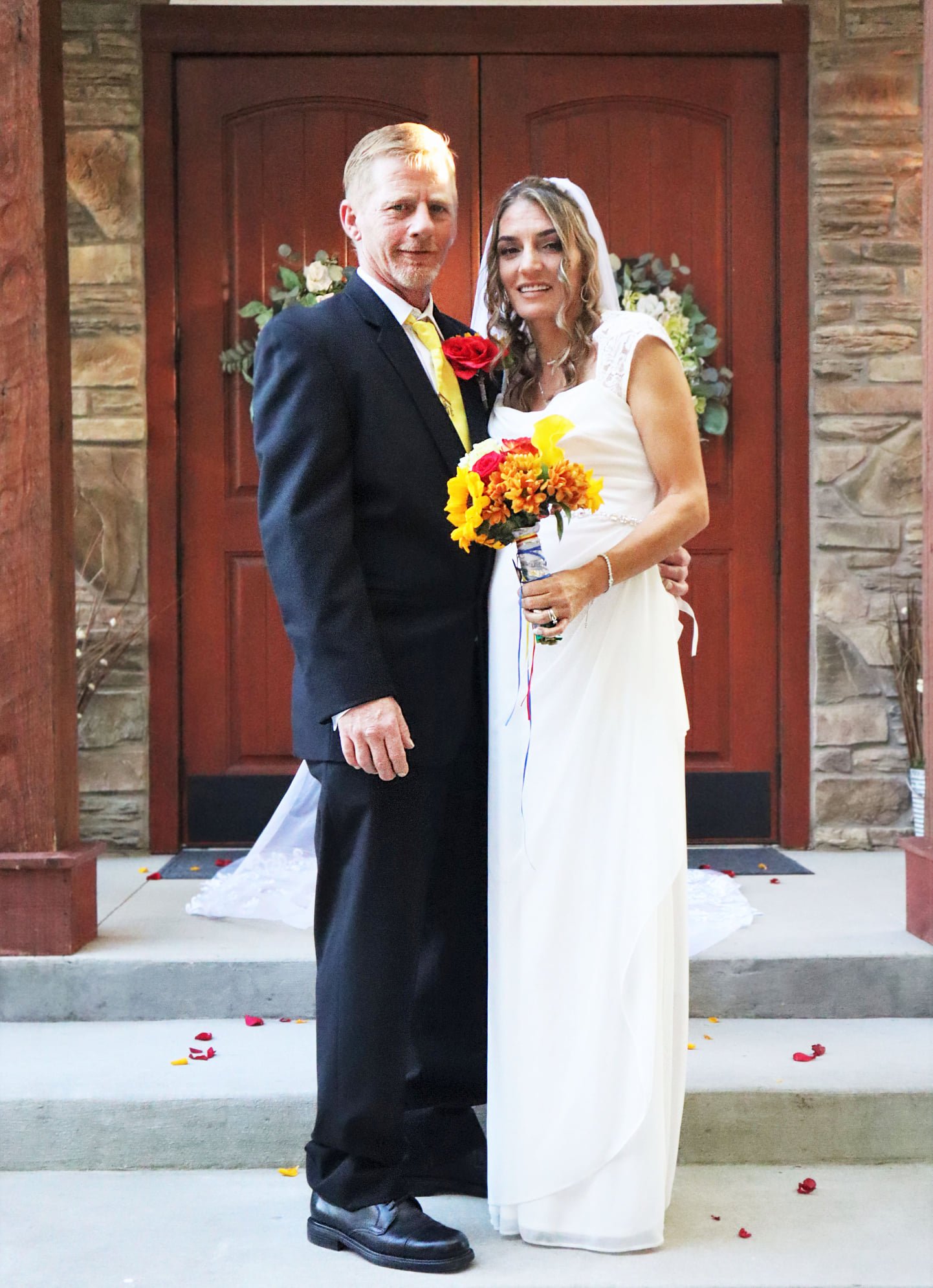 A bride and groom standing together in front of a wooden double door with floral arrangements in the background. The bride holds a bouquet of yellow, orange, and red flowers, and the groom wears a dark suit with a yellow tie and a red rose boutonniere.