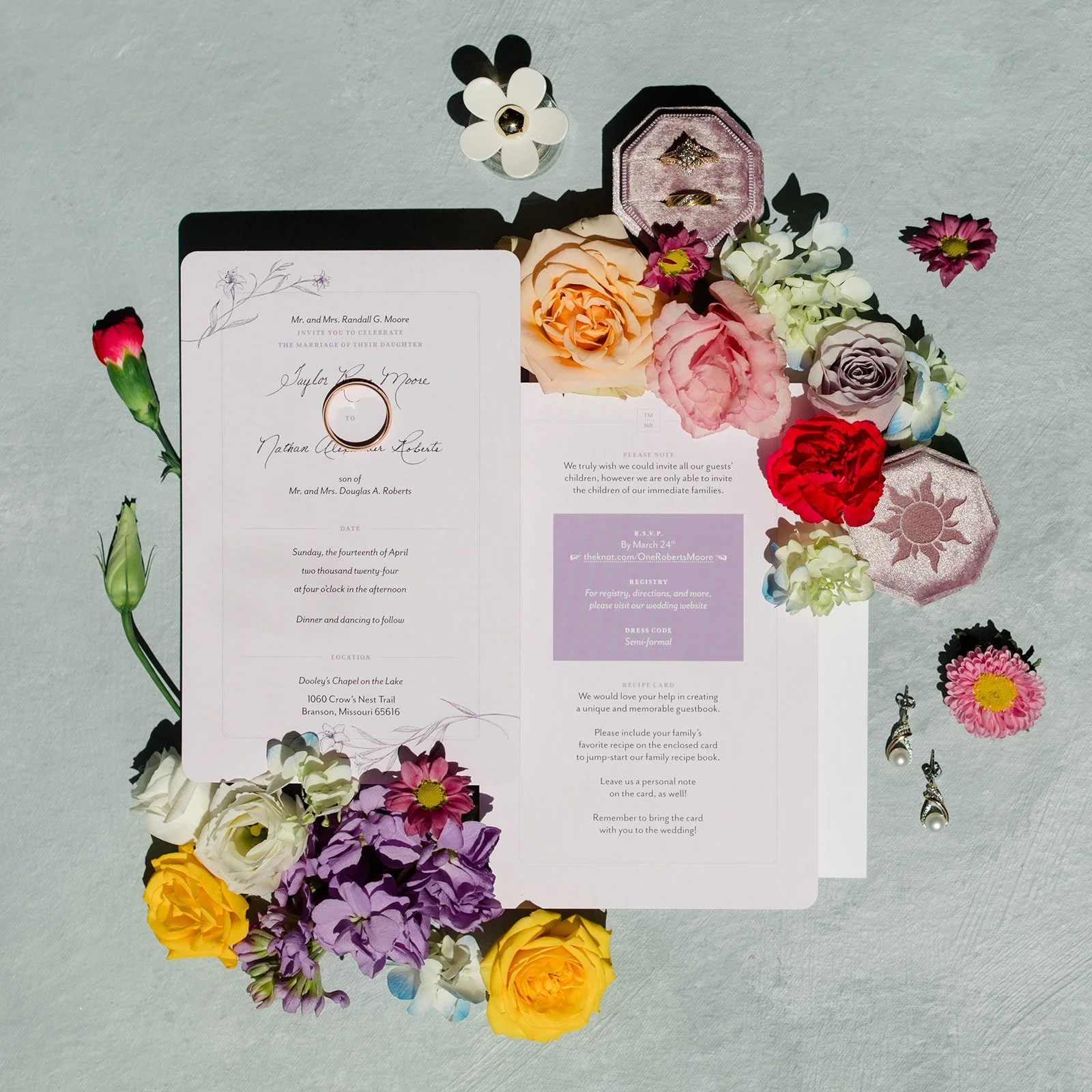 Wedding invitation surrounded by colorful flowers, with rings in a pink ring box and jewelry in a decorative case.