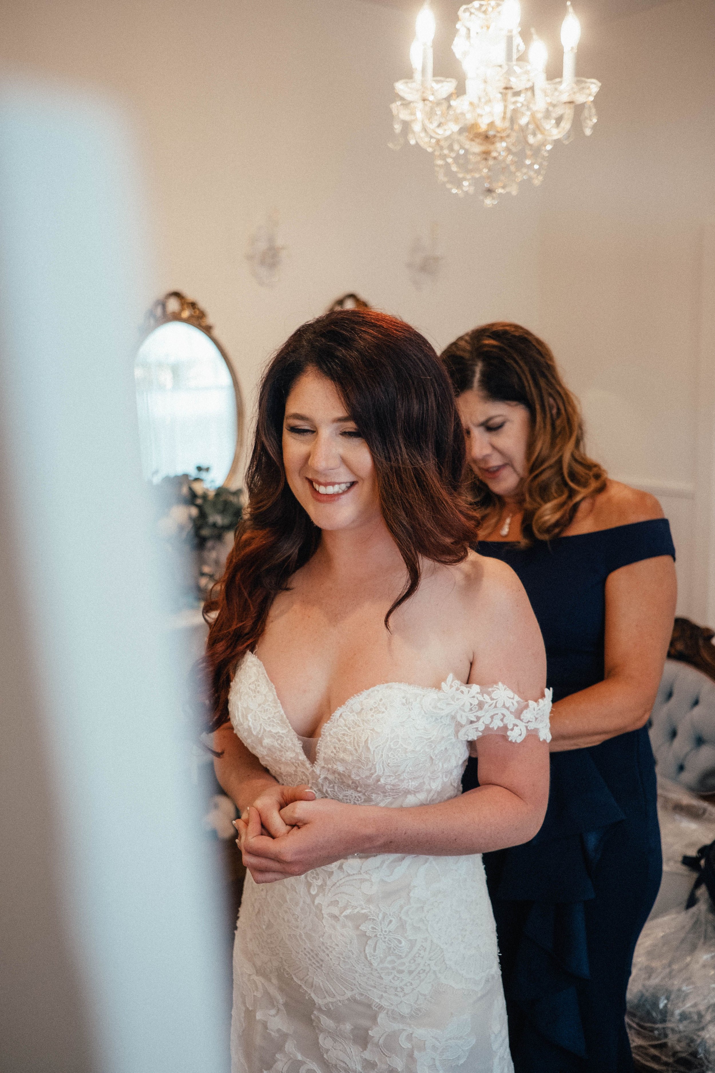 A bride with long, wavy red hair wearing a white lace wedding dress with off-the-shoulder sleeves, smiling with closed eyes, as she stands in a room decorated with a chandelier, mirrors, and floral arrangements. An older woman in a dark blue dress is