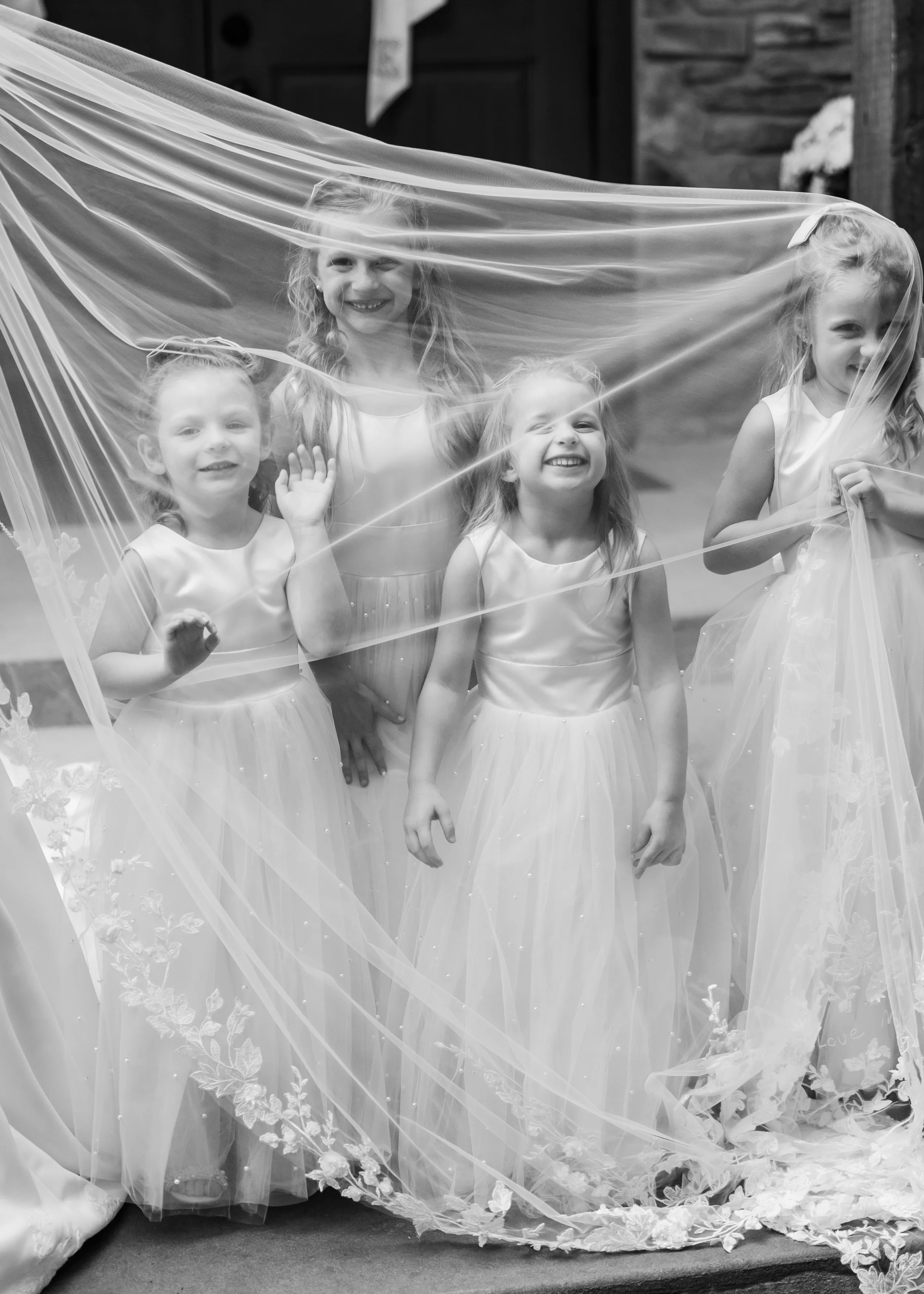 Four young girls in white dresses standing behind a transparent veil with floral embroidery, smiling and waving.