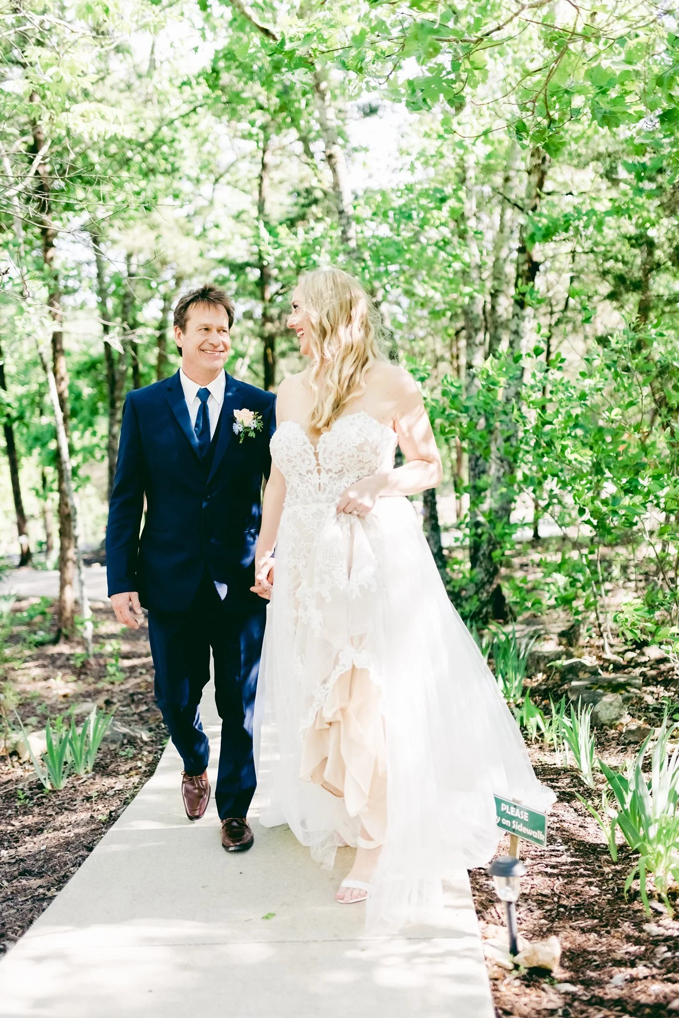 A bride and groom walking on a sidewalk in a park surrounded by trees and greenery, smiling at each other.