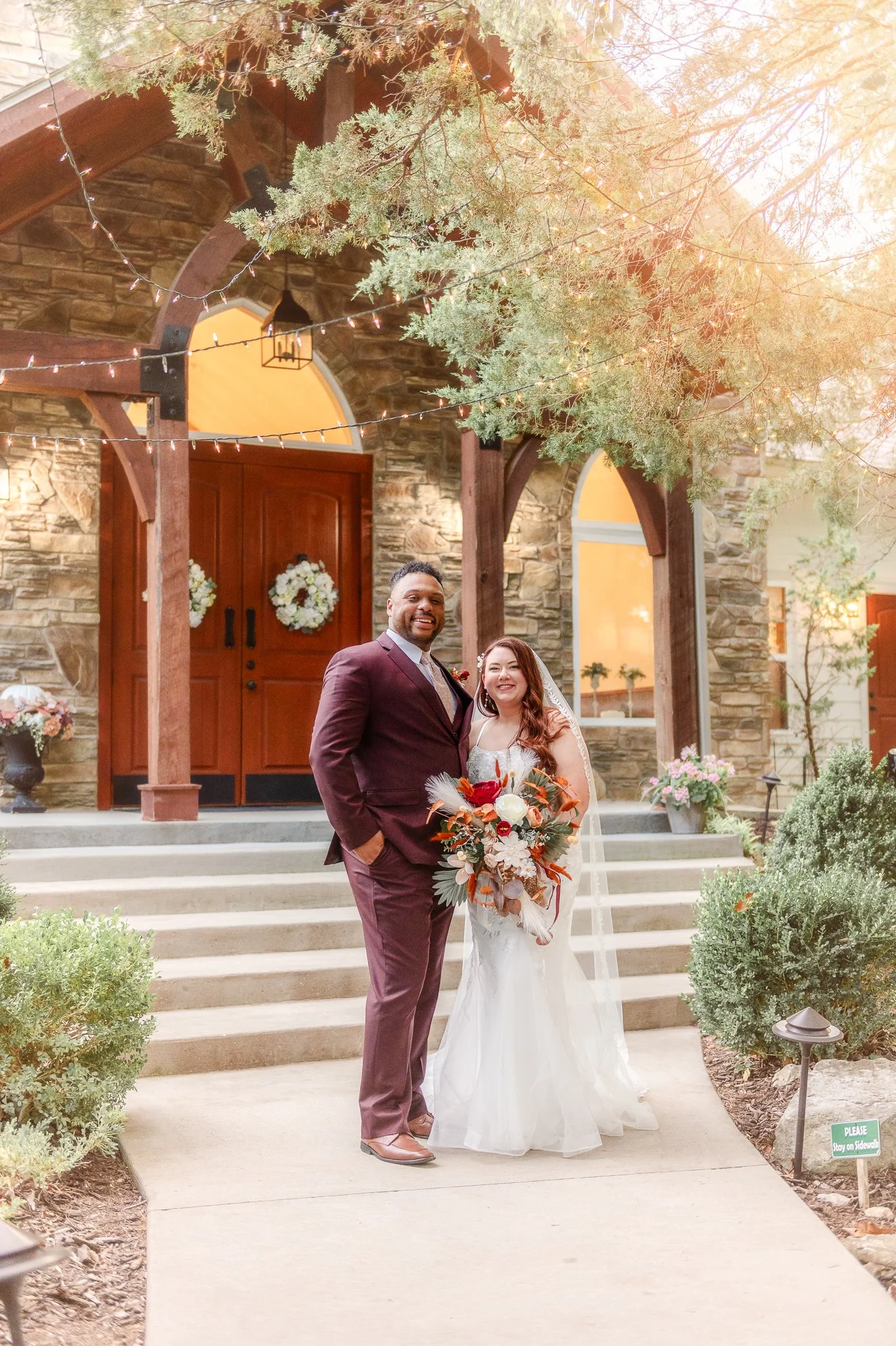 A newlywed couple standing on the steps of a stone building decorated for a wedding, with trees and string lights overhead. The bride is holding a bouquet, and both are smiling.