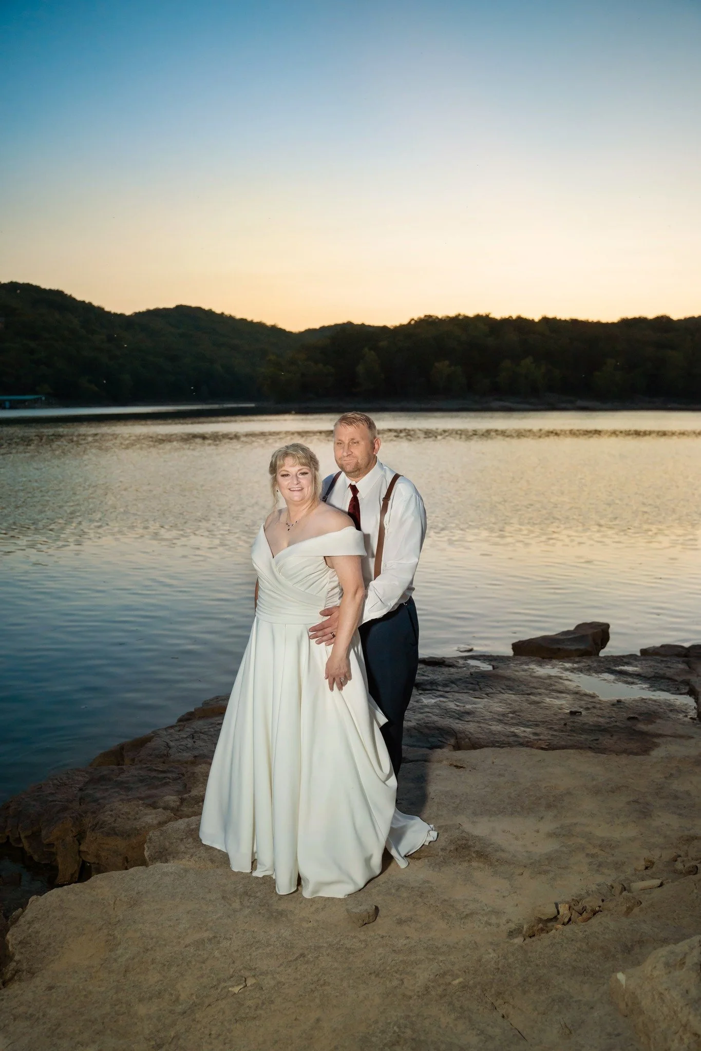 A newlywed couple standing on rocks by the water during sunset, with trees and hills in the background.