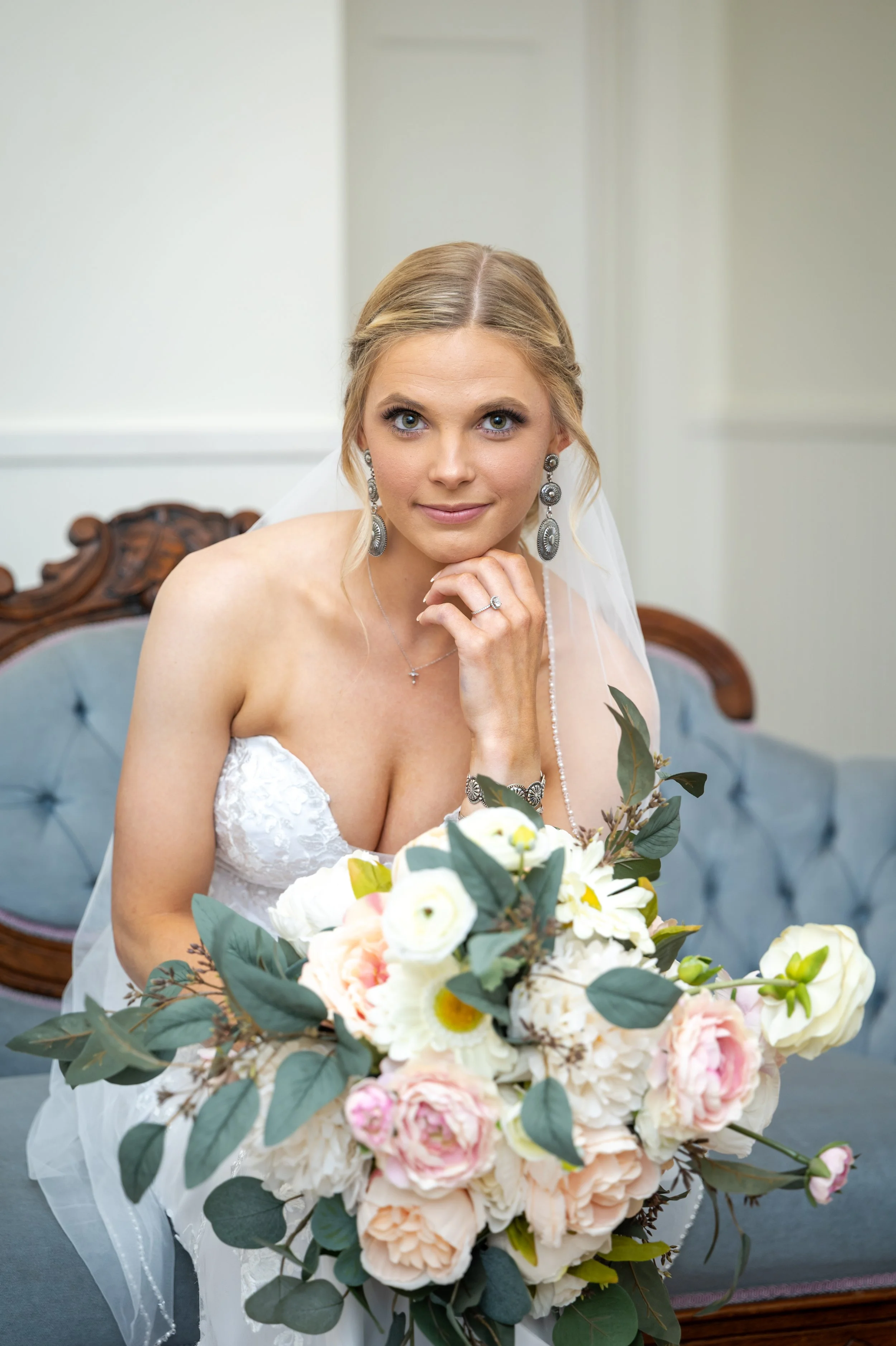 A bride with blonde hair in an updo, wearing large silver earrings, a white lace strapless wedding dress, and holding a bouquet of white and pink flowers with greenery, sitting on a vintage wooden bench in a simple indoor setting.