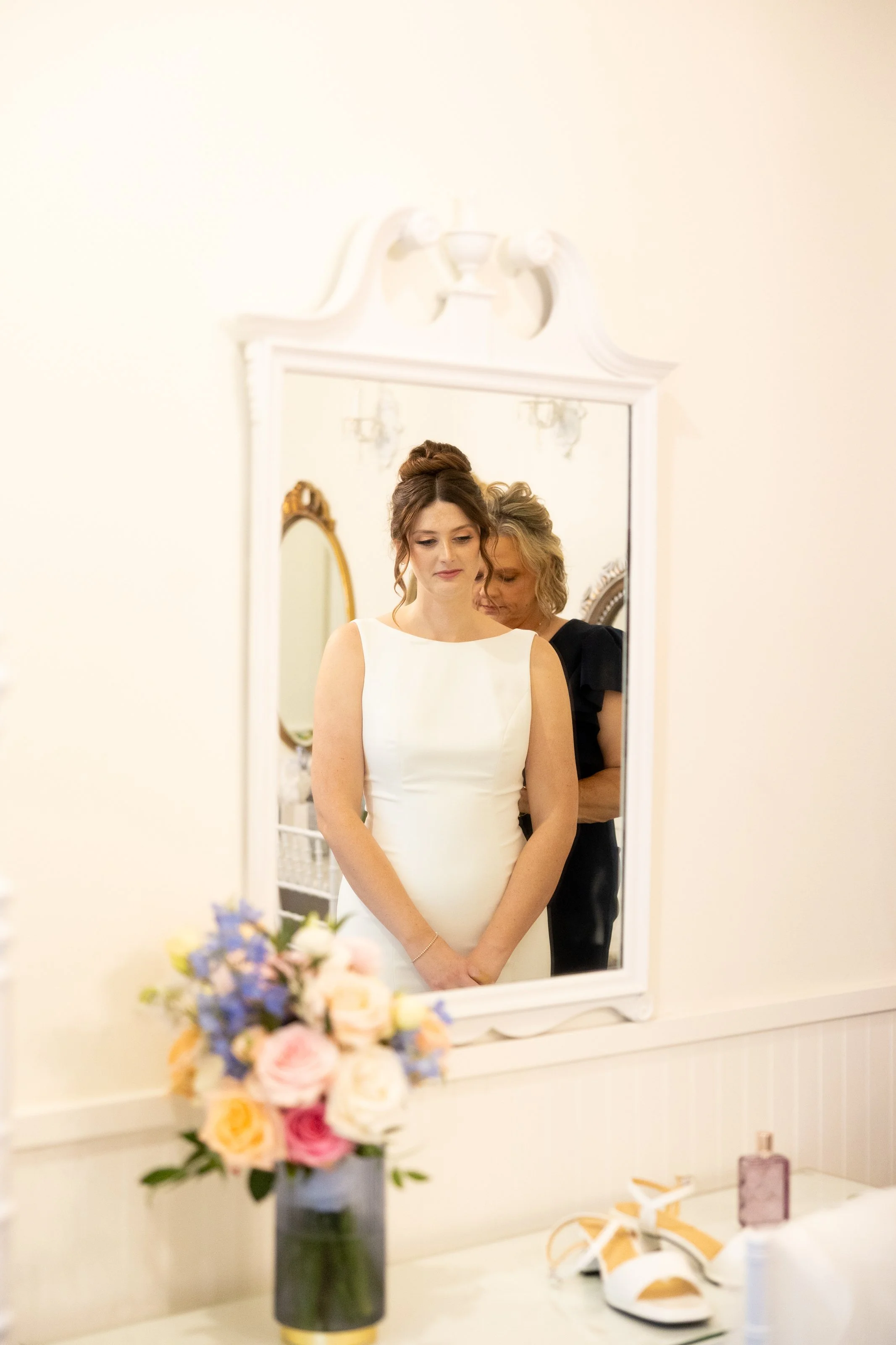A bride in a white dress is looking into a mirror with a woman helping her. The scene is decorated with a floral bouquet on the table and some gift boxes and accessories nearby.