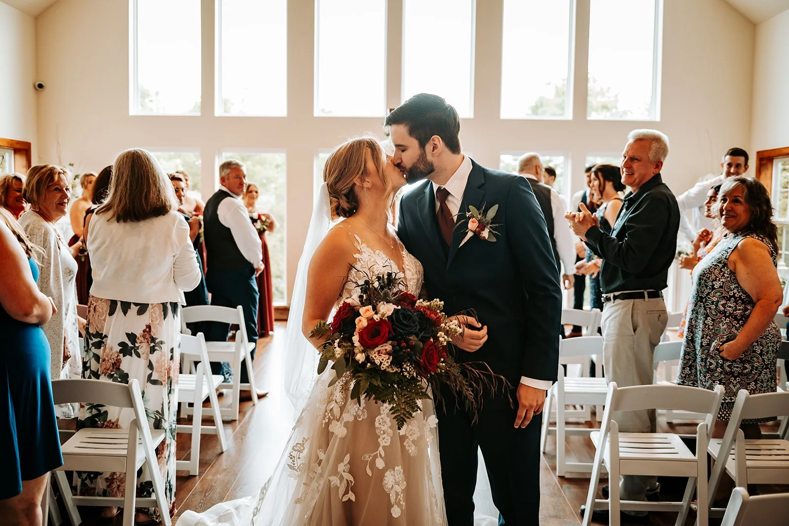 A newlywed couple shares a kiss during their wedding ceremony in a bright indoor venue with guests clapping and smiling in the background.