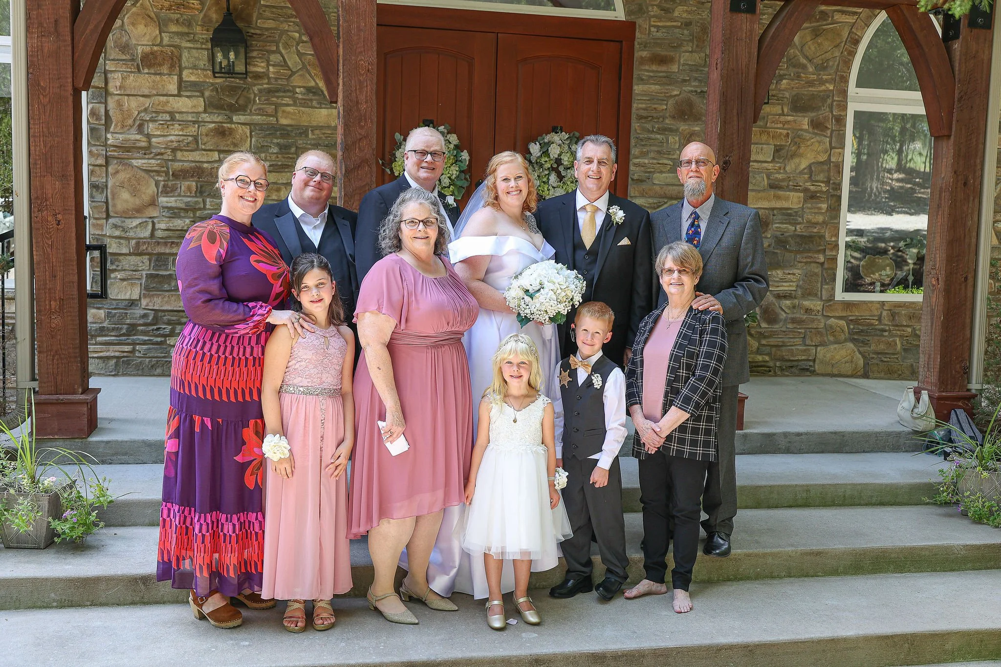 A group of twelve people, including a bride in a white wedding dress holding a bouquet of white flowers, and a groom in a black suit with a gold tie, standing in front of a stone building with a wooden door and floral decorations. They are celebratin
