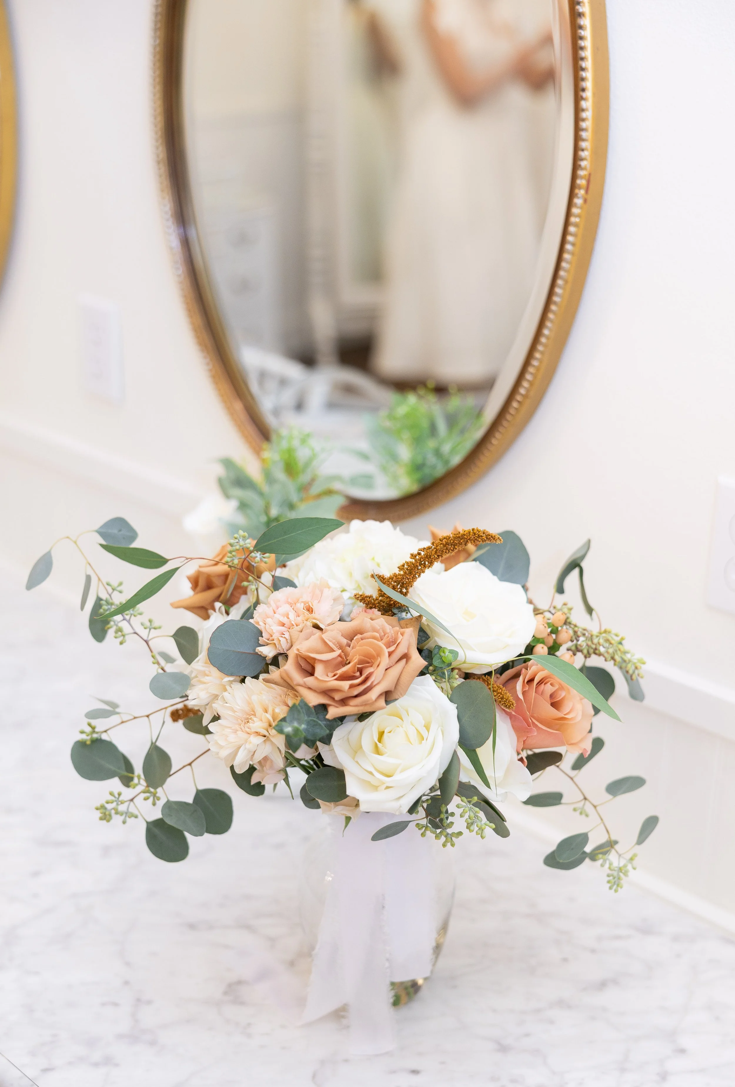 A bouquet of white, cream, peach, and blush pink flowers with greenery in a white vase on a marble surface, with a mirror in the background.