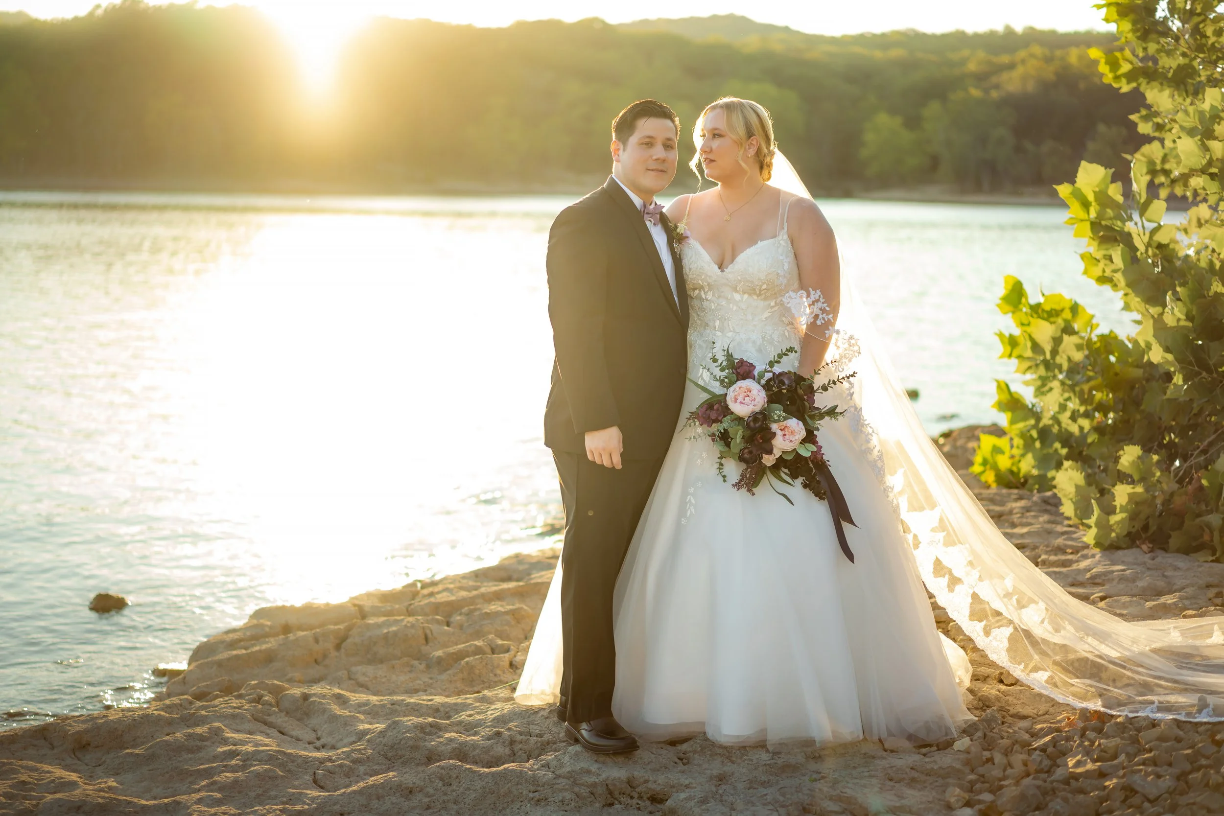 A bride and groom standing on a rocky beach near a body of water during sunset. The bride is wearing a white wedding gown with lace details and a veil, holding a bouquet of pink and purple flowers. The groom is dressed in a black tuxedo with a bow ti