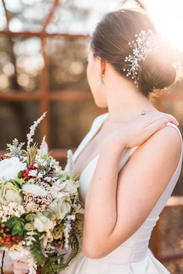 A bride with an elegant updo hairstyle decorated with a sparkling hairpiece, wearing a sleeveless white wedding gown, holding a bouquet of flowers, outdoors on a sunny day.