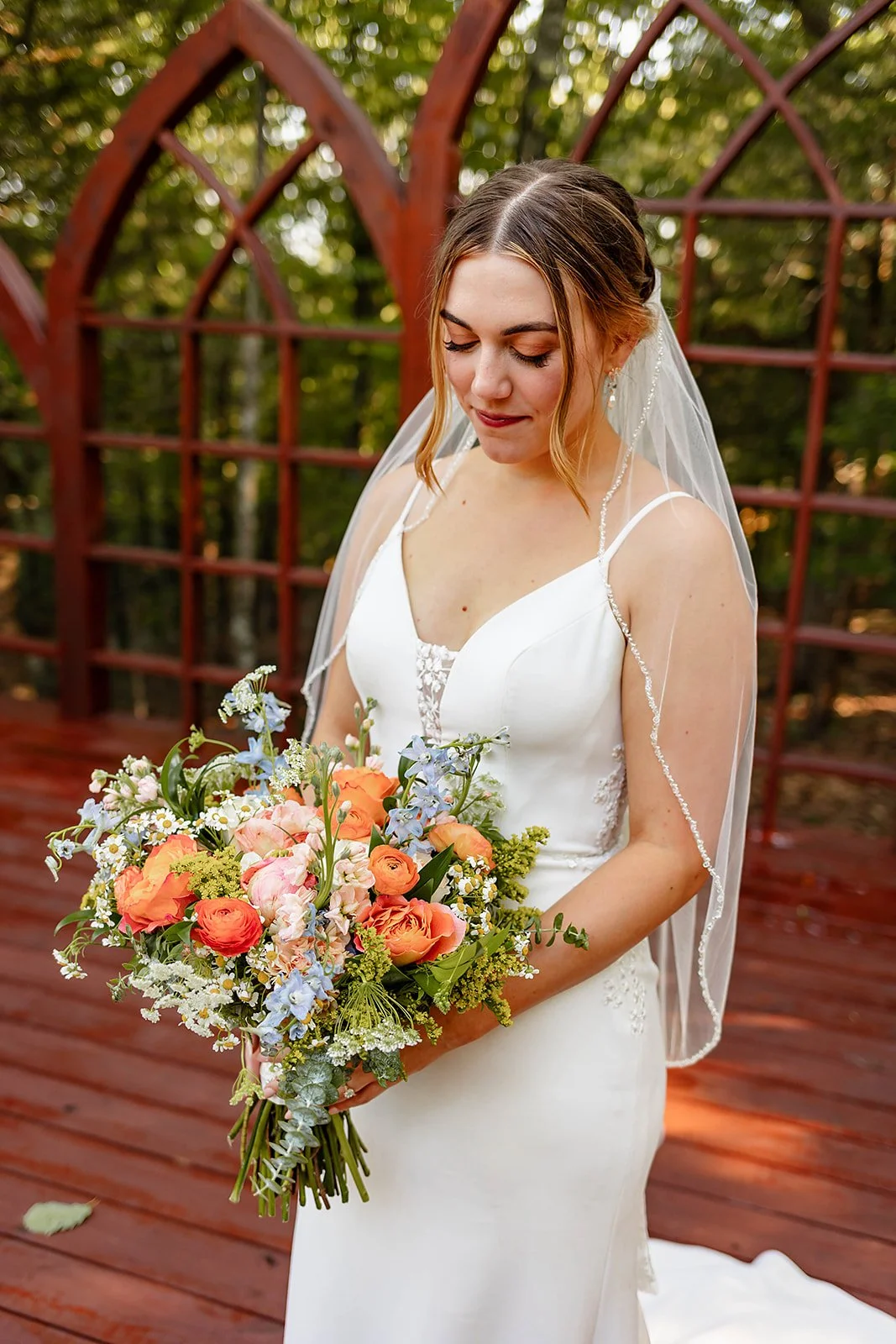 A bride in a white wedding dress holding a colorful bouquet of flowers, standing outdoors on a wooden deck with a red glass-paneled structure and green trees in the background.