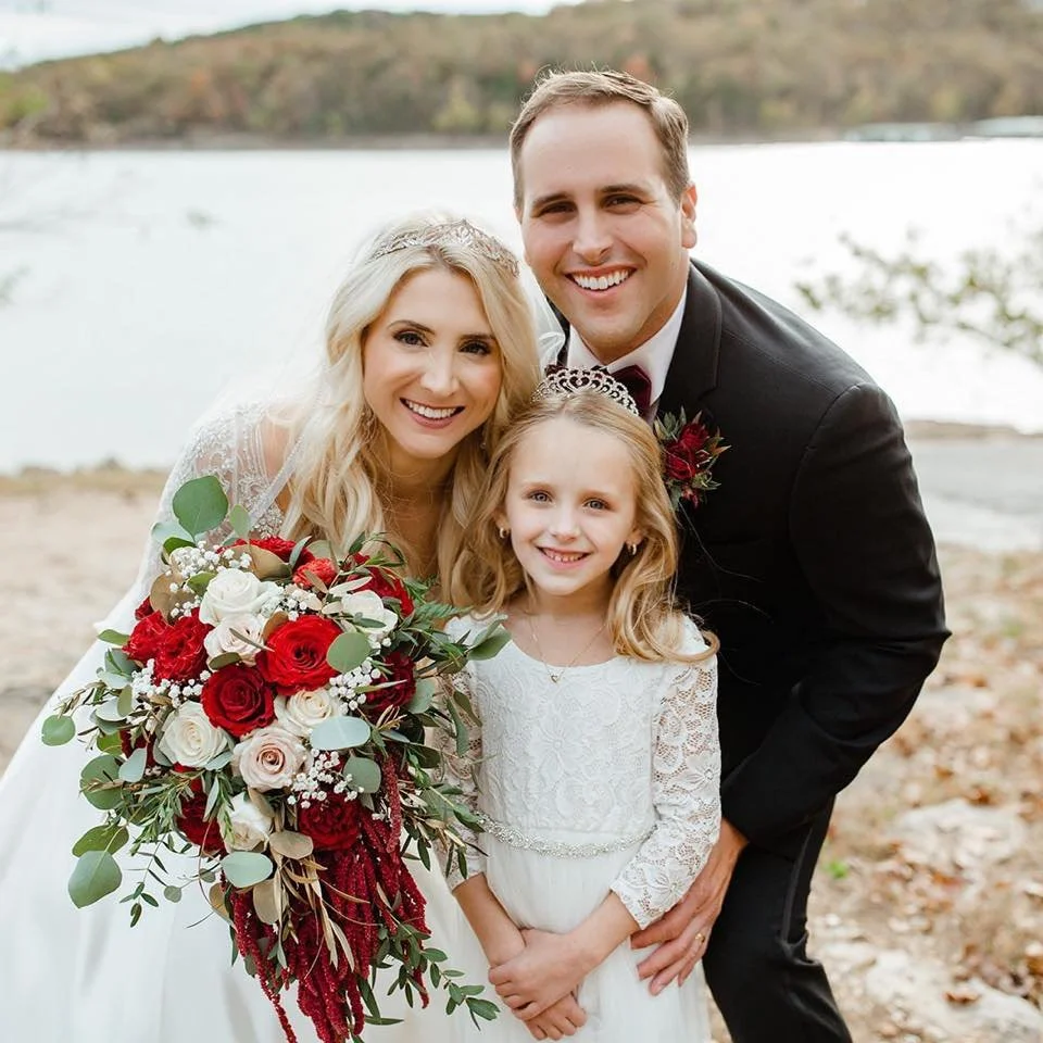 A family of three, a bride, groom, and young girl, smiling outdoors near a body of water with trees in the background. The bride holds a bouquet of red and white flowers, and the girl wears a white lace dress while the groom is dressed in a black suit.