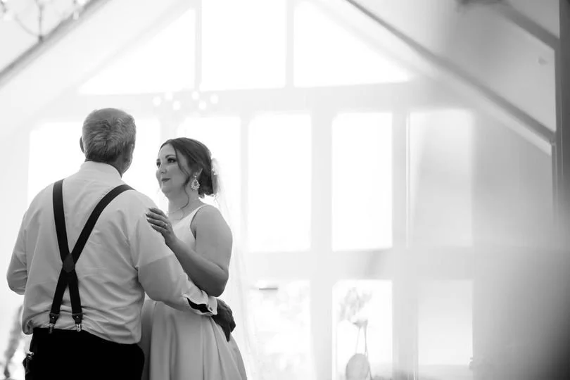 A bride and an older man, likely her father, dancing together at a wedding reception in a bright room with large windows.