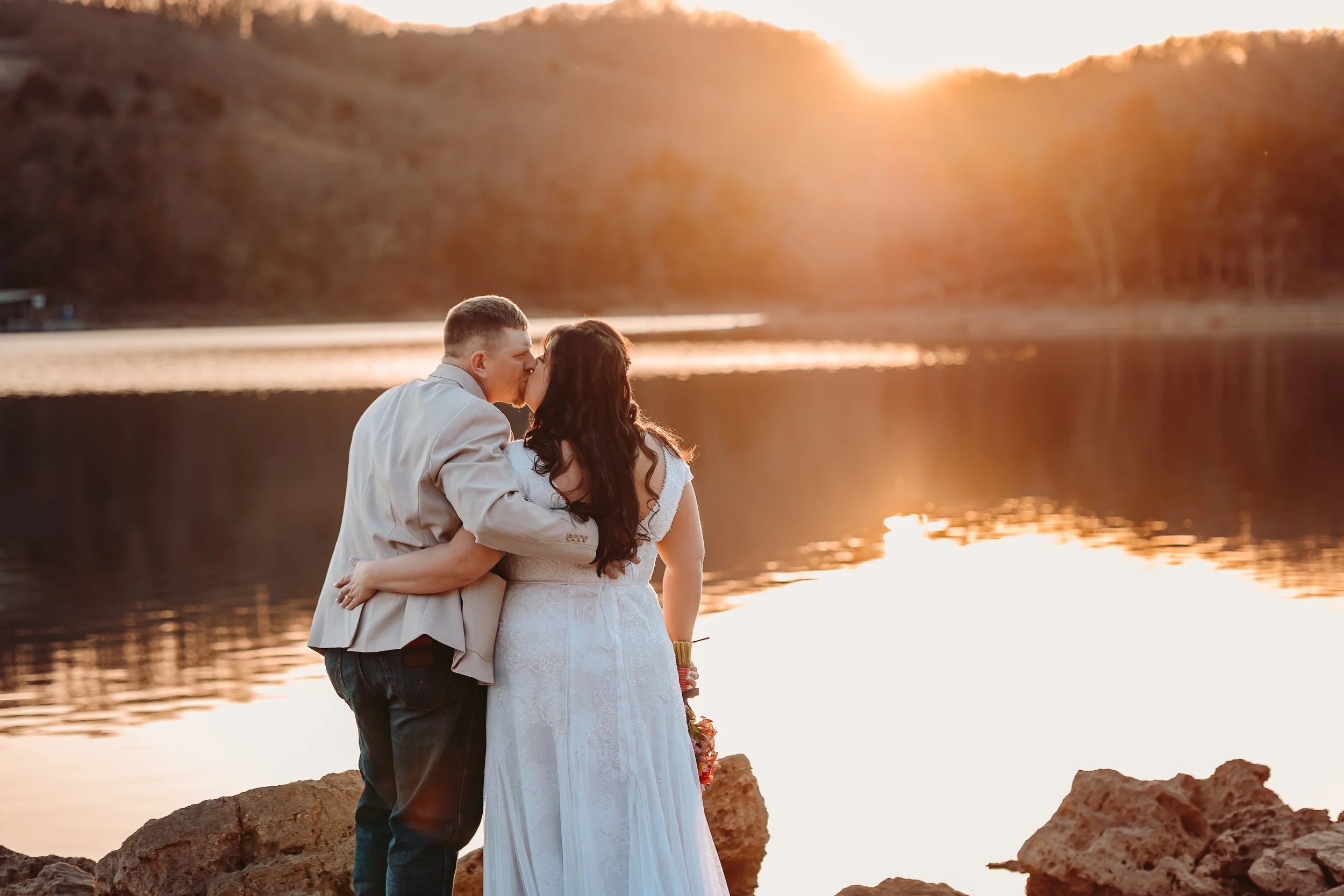 A couple dressed in wedding attire sharing a kiss near a lake at sunset. The man is in a light-colored suit jacket and dark pants, and the woman is in a white wedding dress with lace details, holding a bouquet. They are standing on rocky terrain, with calm water and a hillside in the background.