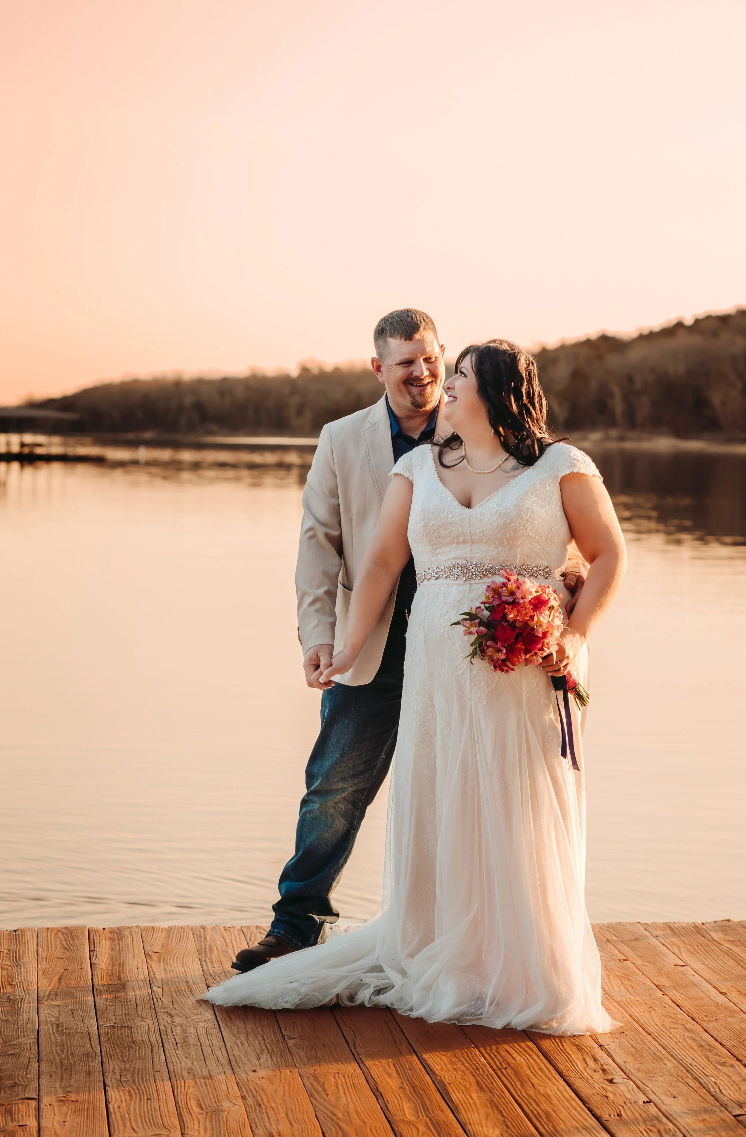A newlywed couple standing on a wooden dock by a lake at sunset, smiling at each other. The bride is holding a bouquet of pink and orange flowers, and the groom is dressed in a light blazer and jeans.