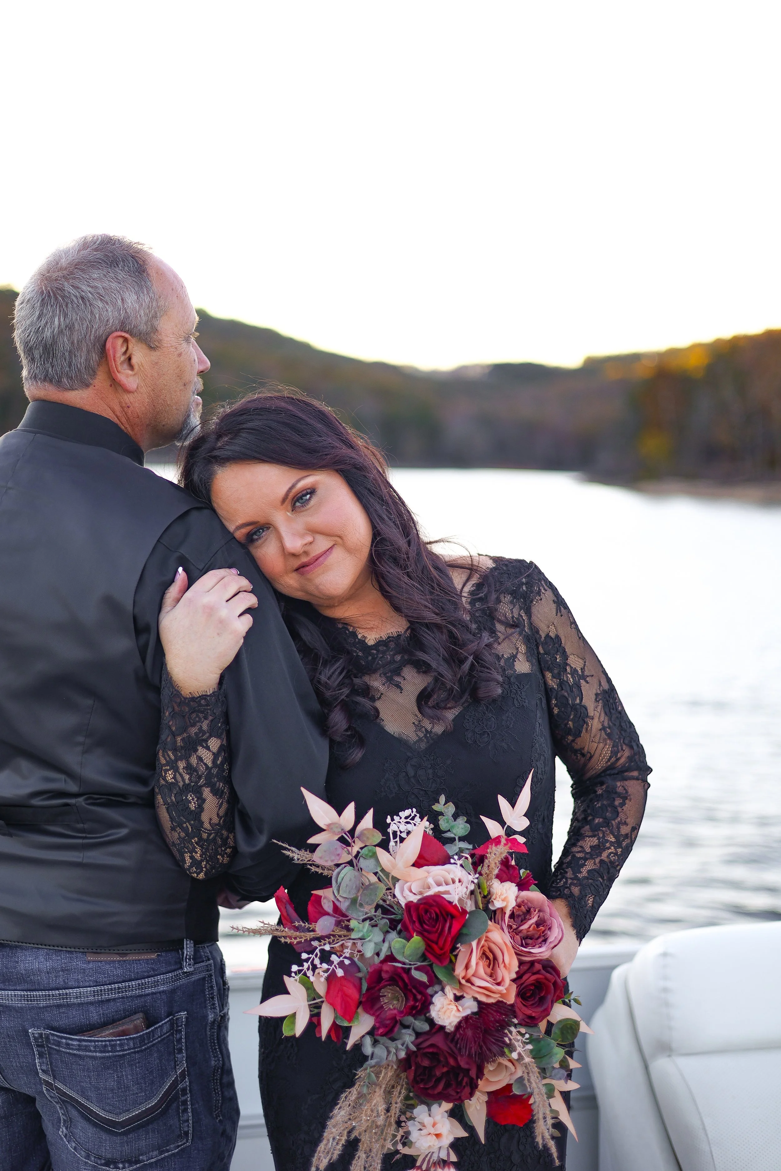 A woman in a black lace dress holding a large bouquet of pink, red, and burgundy flowers, with a man in a black jacket nearby, outdoors near a body of water with trees in the background.