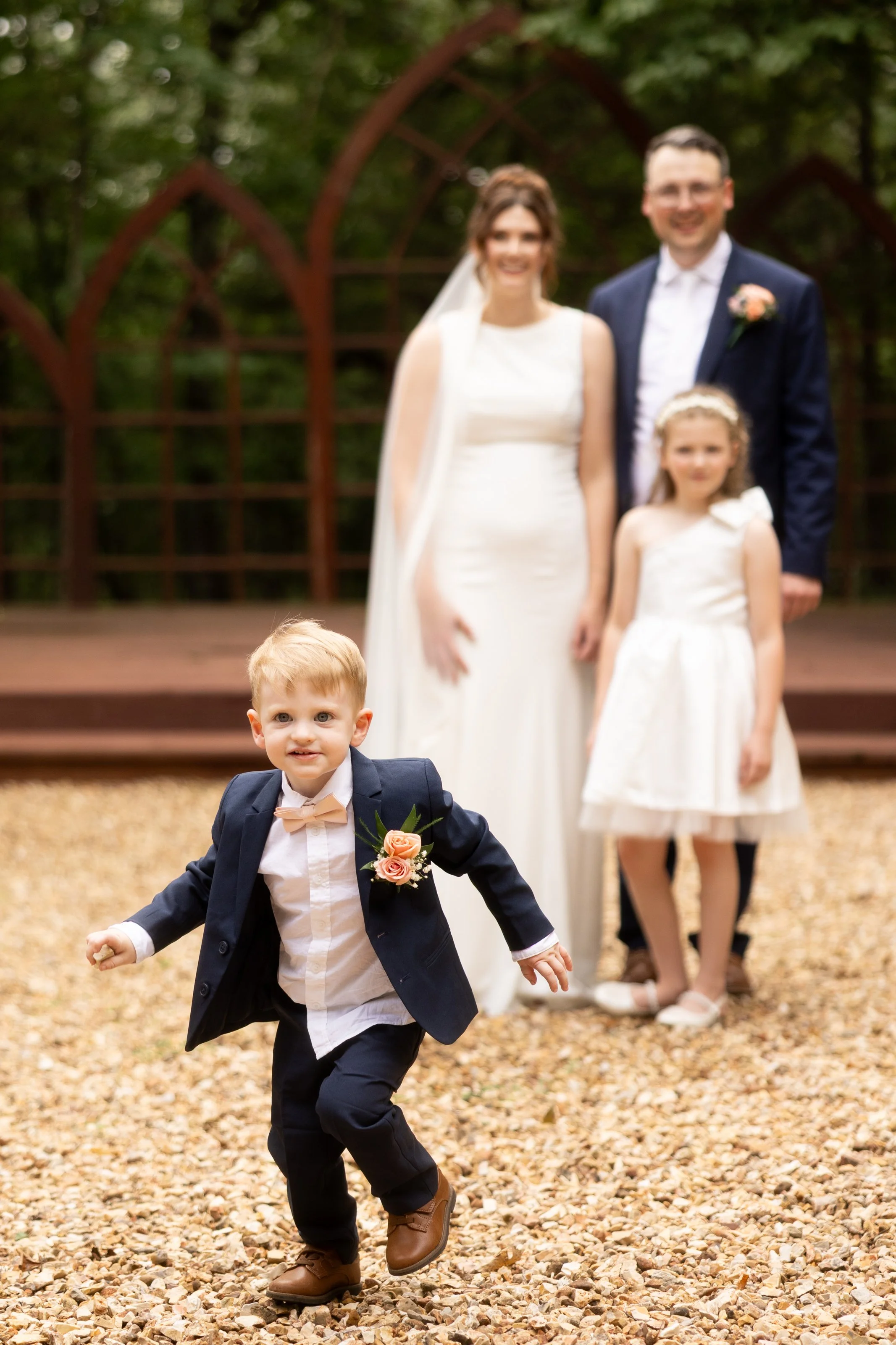 Young boy in a tuxedo running toward the camera with a joyful expression at a wedding reception, with a blurred bride, groom, and flower girl in the background on a gravel outdoor setting.