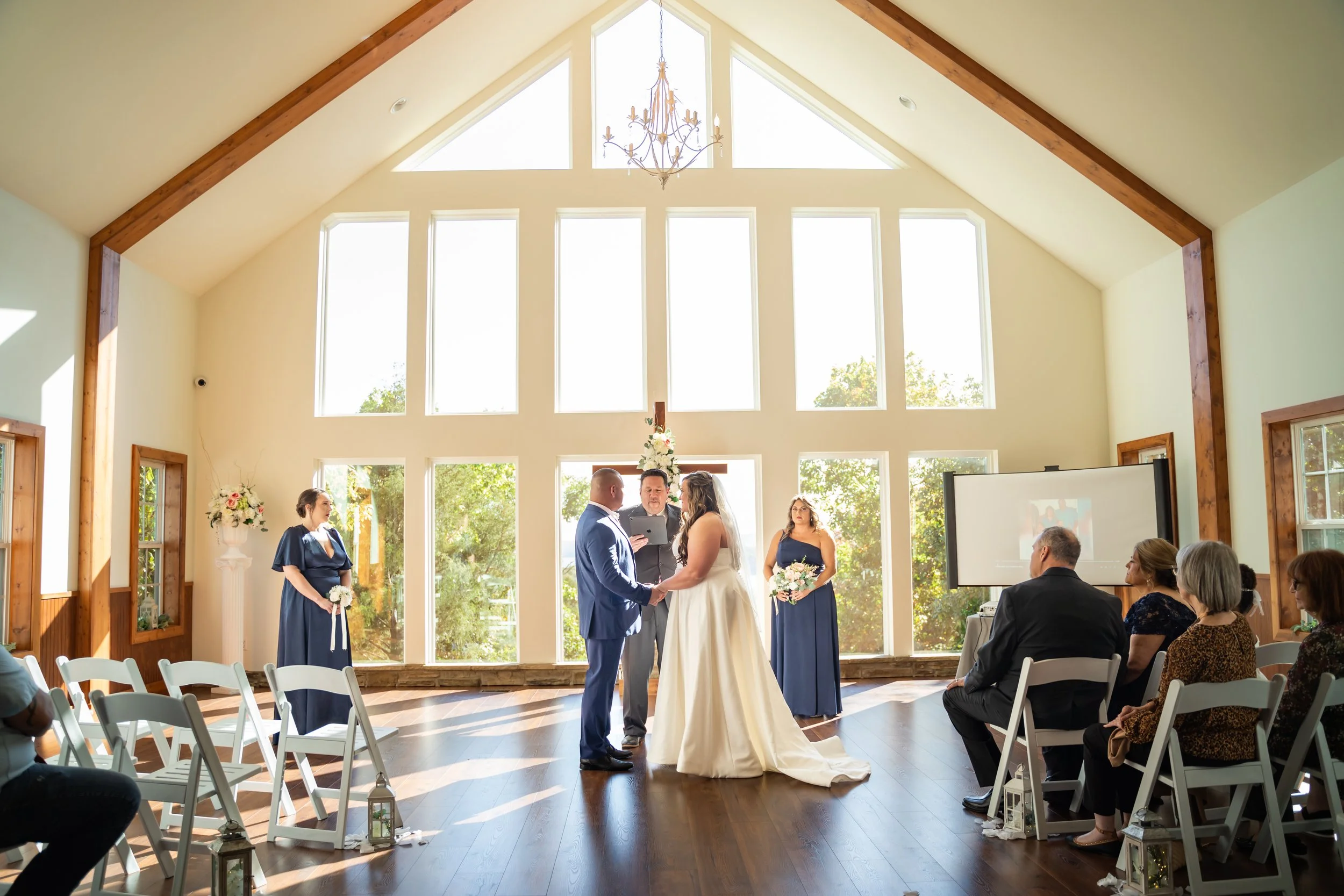 A wedding ceremony in a bright, spacious room with large windows, wooden accents, and a chandelier. The bride and groom are standing in front, holding hands, exchanging vows, while officiant and bridesmaids stand nearby. Guests are seated watching the ceremony.