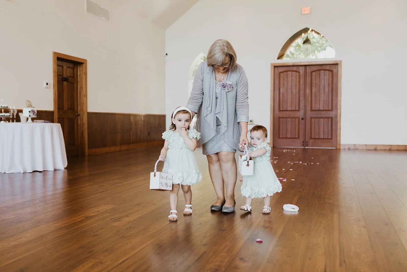 An older woman, possibly a grandmother, walking with two young girls in white dresses inside a spacious room with wooden floors and large doors. The girls are holding small baskets and have flower girl outfits, indicating a wedding setting. The woman