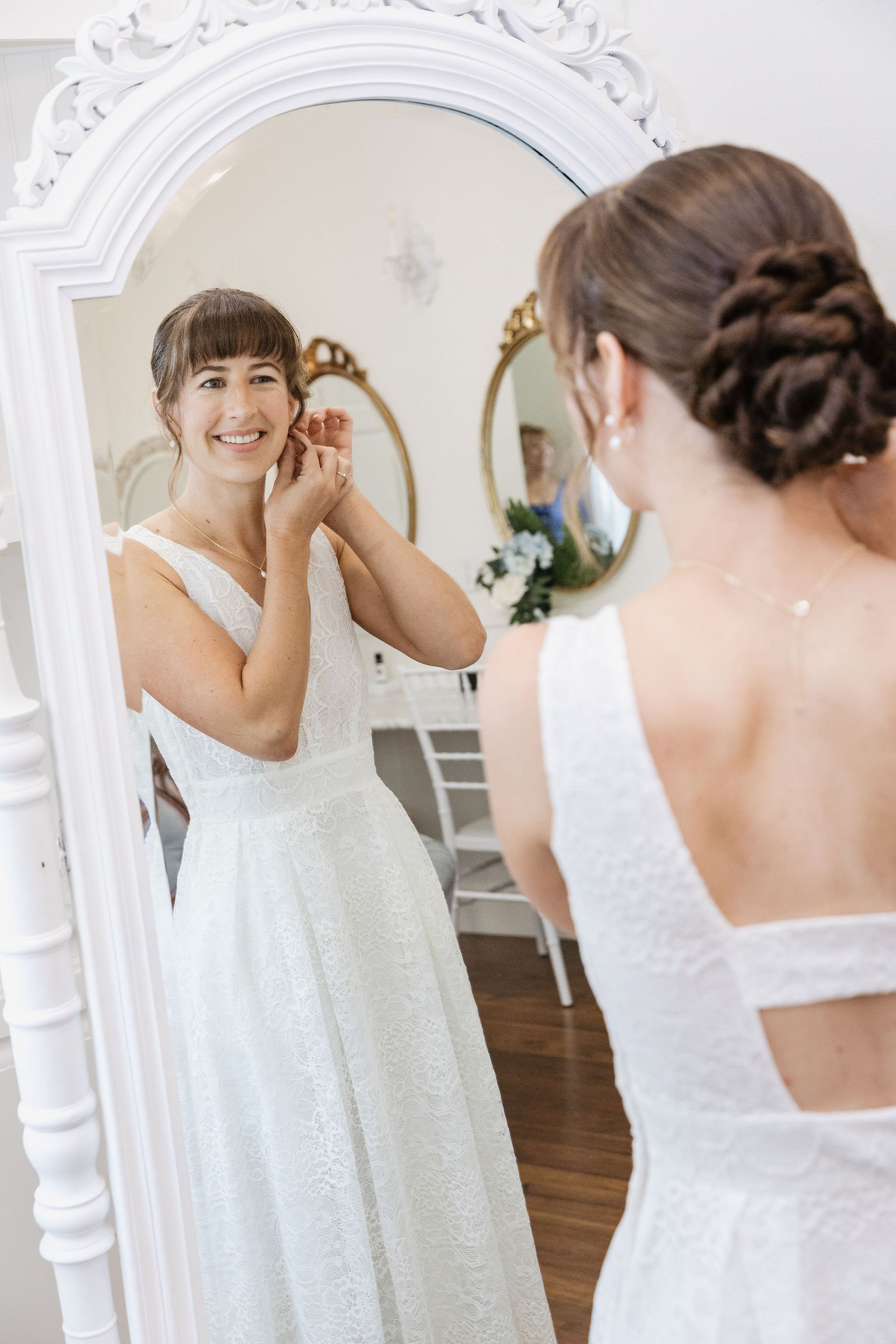 A woman in a white lace dress is smiling at herself in an ornate white-framed mirror while adjusting her earring.