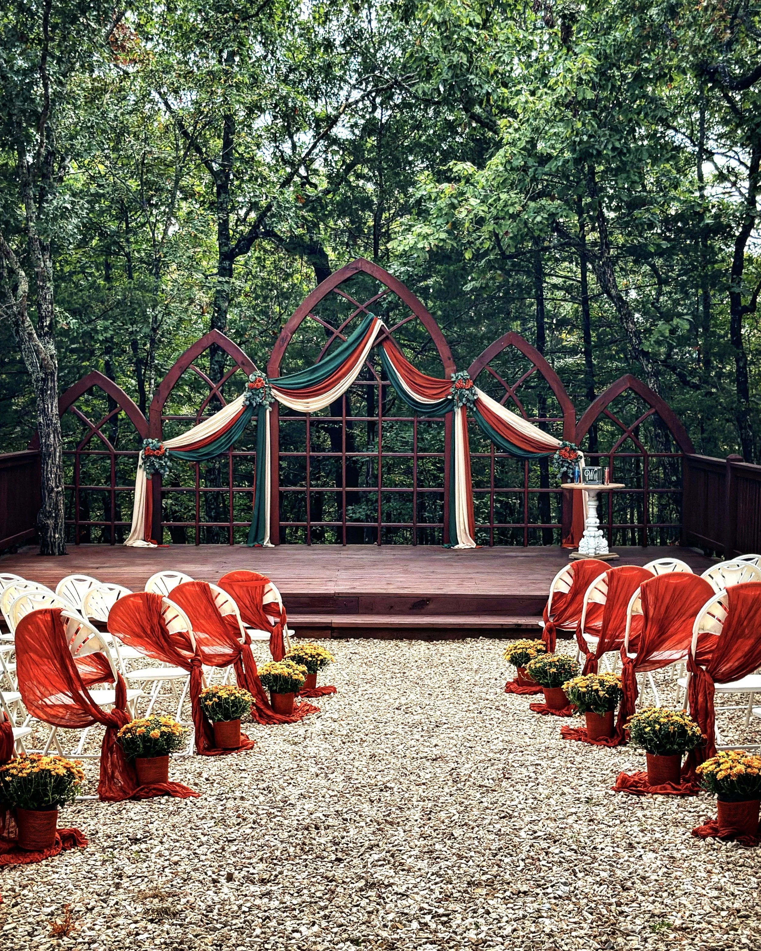 Exterior of a wedding ceremony area set up outdoors in a wooded area with trees in the background. The setup includes a wooden arch decorated with draped fabric in red, cream, and green, supported by flower arrangements. Rows of white chairs with red