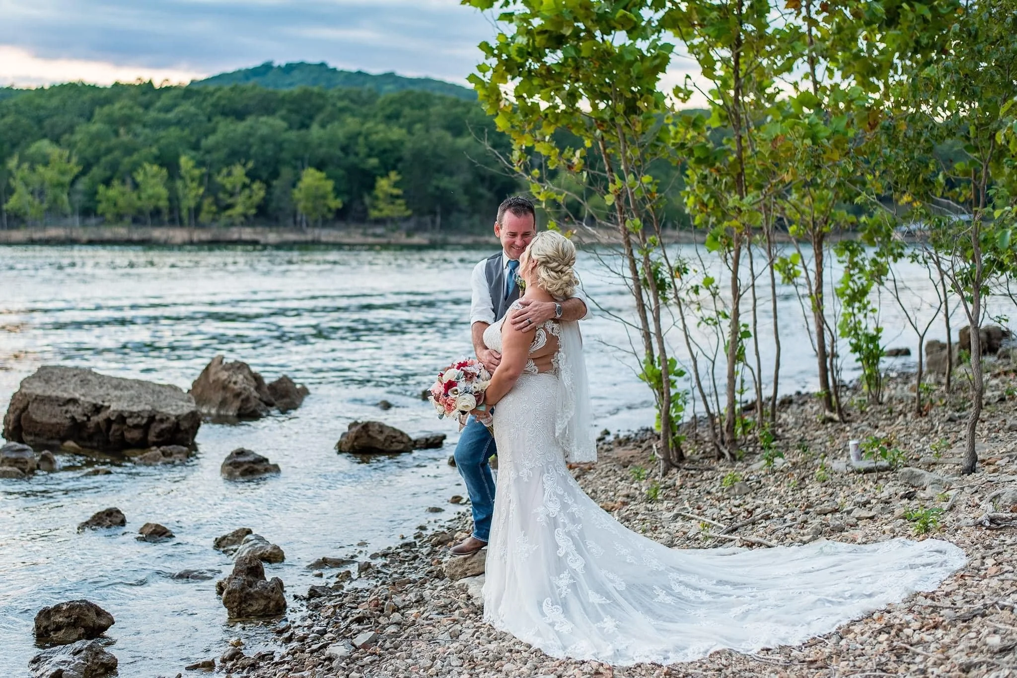 A bride and groom embrace by a river on their wedding day, surrounded by trees and rocks with a hilly forested landscape in the background.