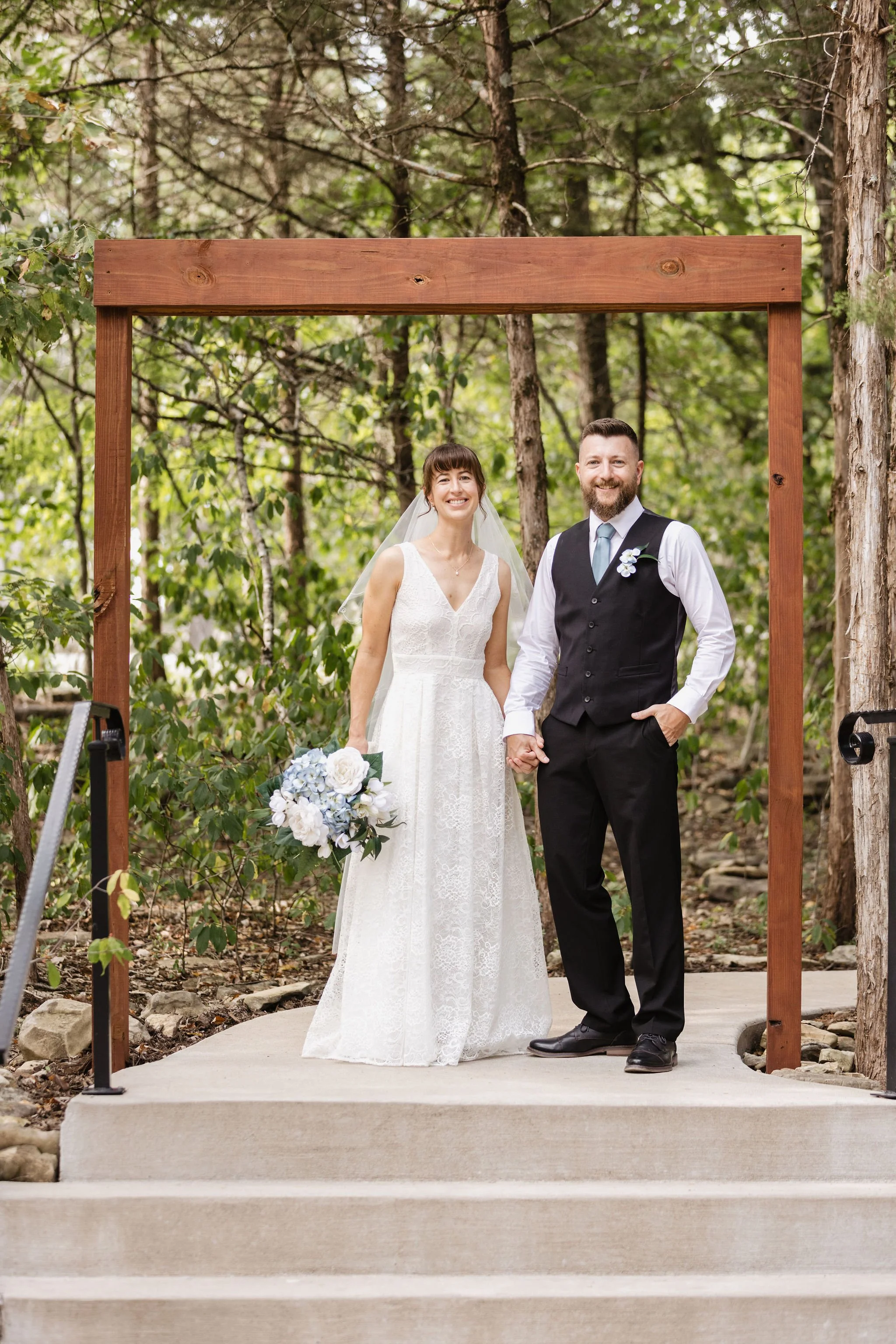 A bride and groom standing hand in hand outdoors during a wedding ceremony, smiling at the camera, under a wooden arch with a forest background.