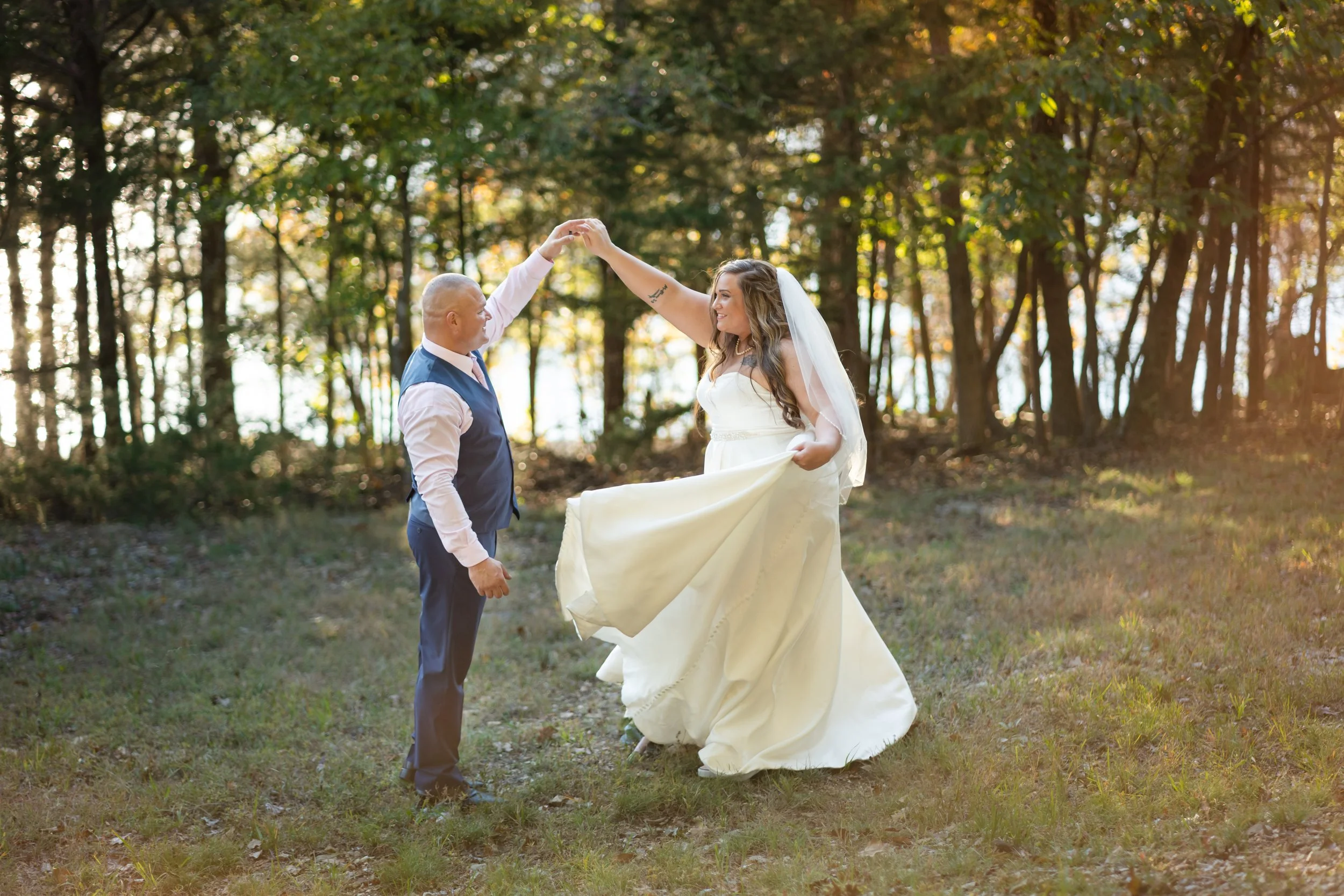 A couple dancing outdoors in a wooded area during fall, with sunlight filtering through the trees. The woman wears a white dress and a veil, while the man is dressed in a vest and tie. They are holding hands and smiling at each other.