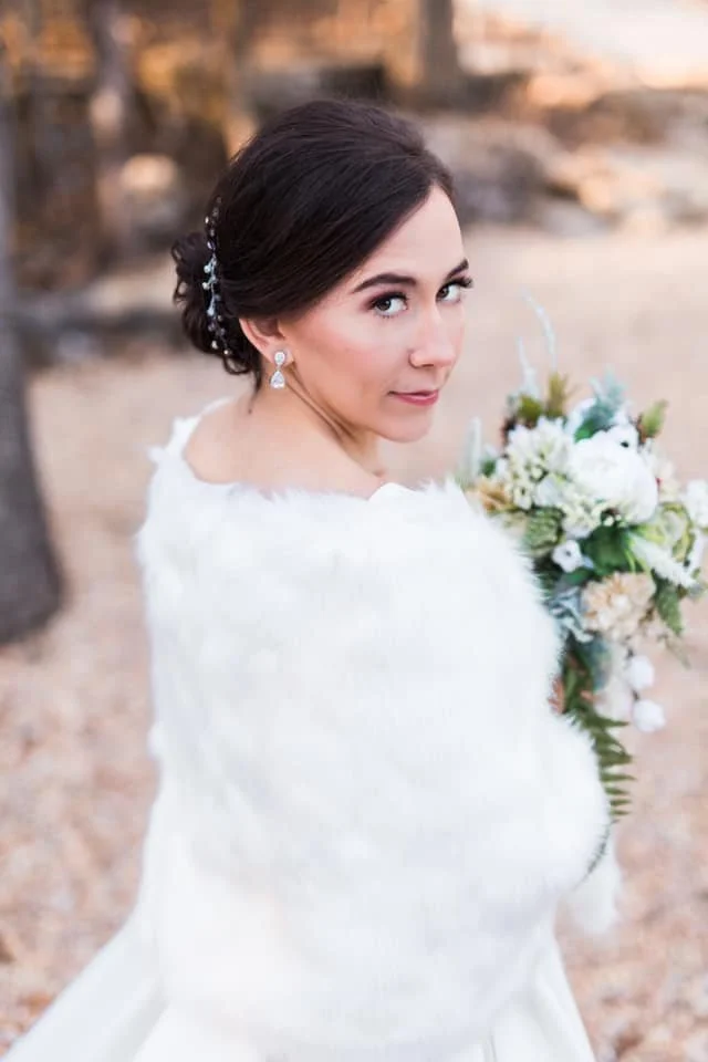 Bride in a white dress holding a bouquet of white and green flowers, looking over her shoulder at the camera, outdoor setting with trees and rocks in the background.