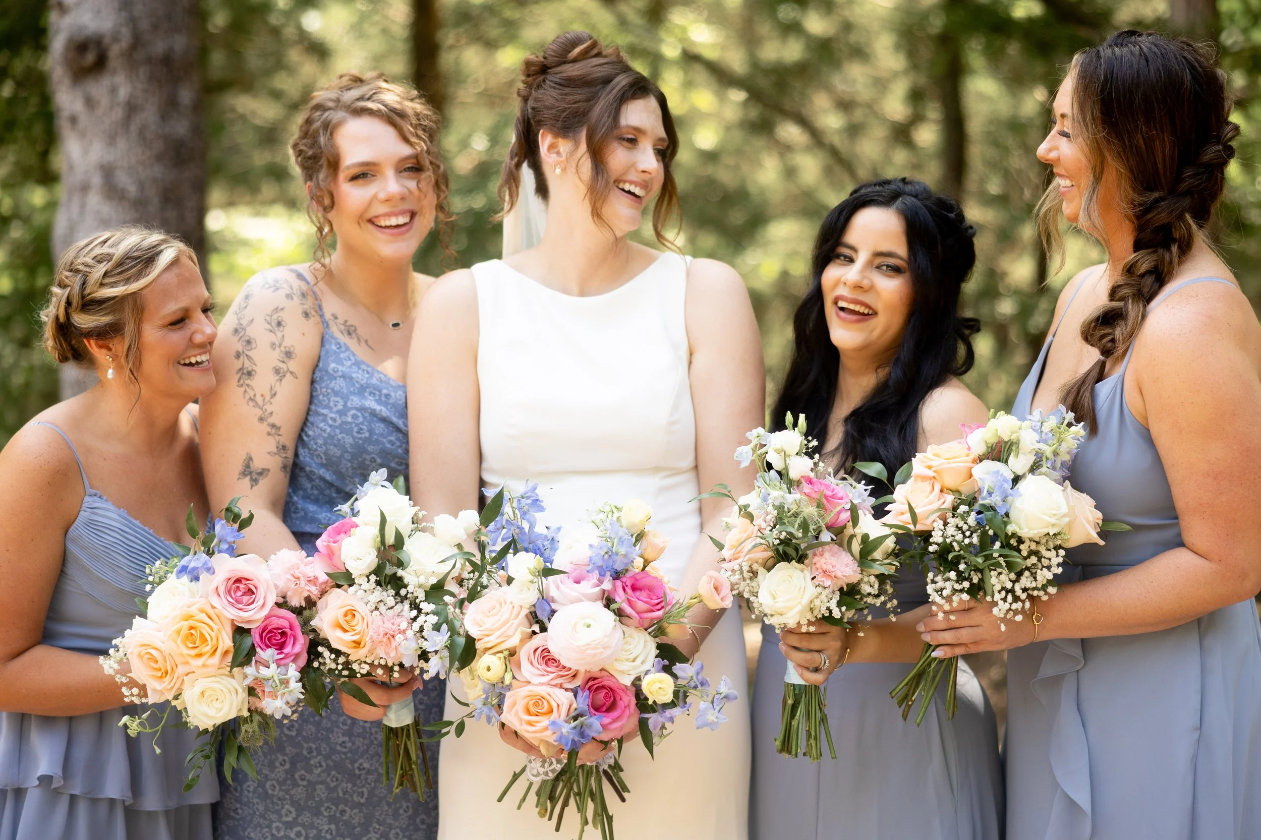A bride and five bridesmaids standing outdoors among trees, each holding a bouquet of colorful flowers, dressed in pastel shades of blue, gray, and white, smiling and celebrating a wedding.