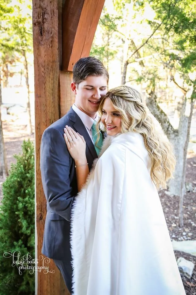 A happy couple standing close together outdoors, smiling, under a wooden structure with trees and sunlight in the background.