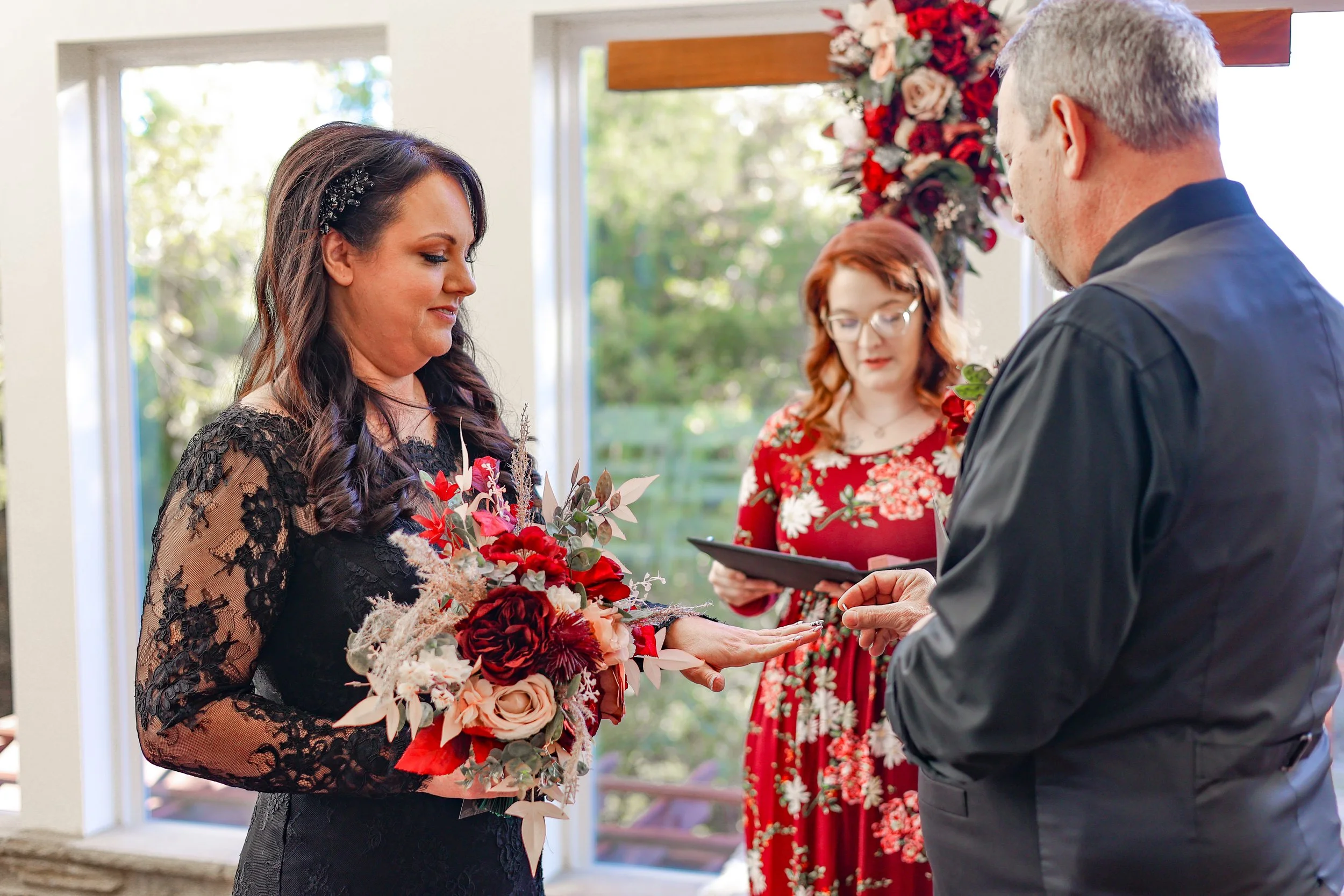 A woman in a black lace dress holds a bouquet of red, pink, and white flowers as she participates in a wedding ceremony. A man in a dark suit appears to place a ring on her finger. A woman in a red floral dress stands behind them, holding a small book or folder. The background includes large windows with greenery outside and a floral arrangement on the wall.