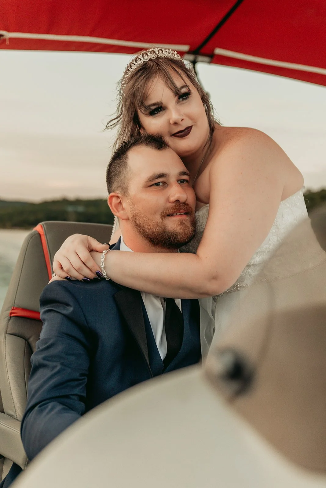 A newlywed couple sitting in a boat, with the bride in a wedding dress and tiara, and the groom in a suit, embracing under a red umbrella during sunset.