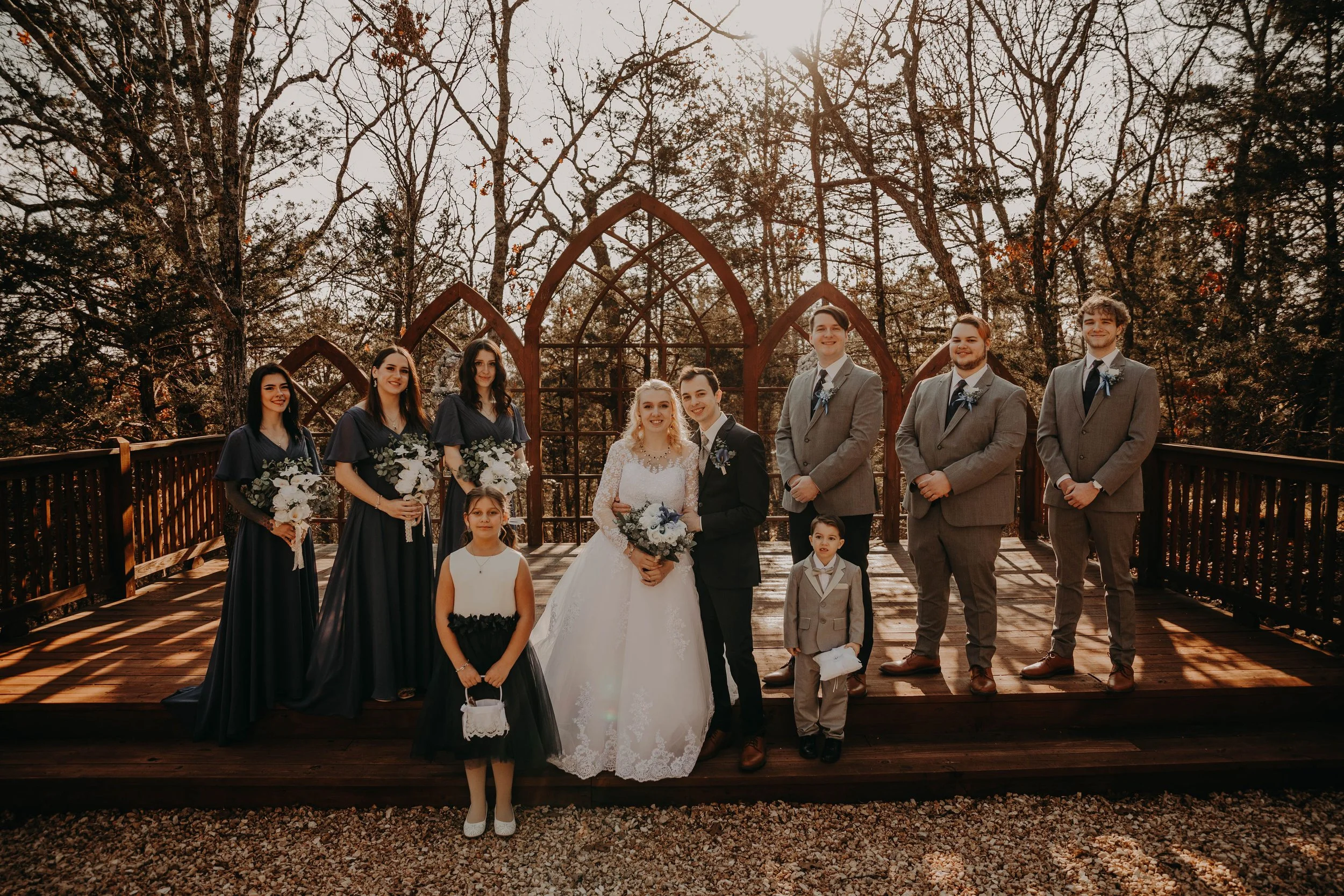 A wedding party gathered outdoors on a wooden deck with a metal arched structure behind them, surrounded by trees with some leaves, during late afternoon sunlight.
