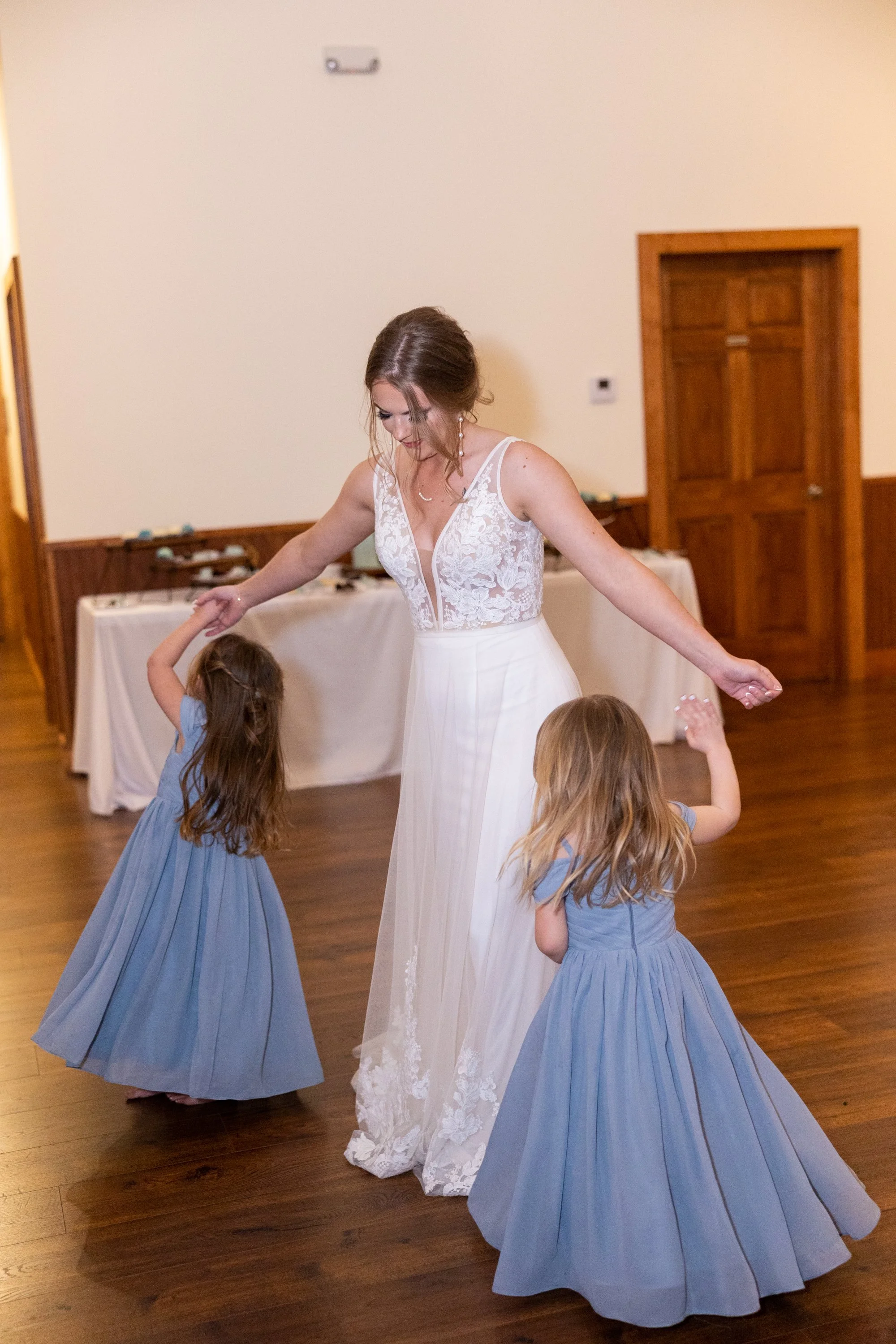 A woman in a white lace wedding dress dancing with three young girls in blue dresses in a room with a wooden floor and cream-colored walls.