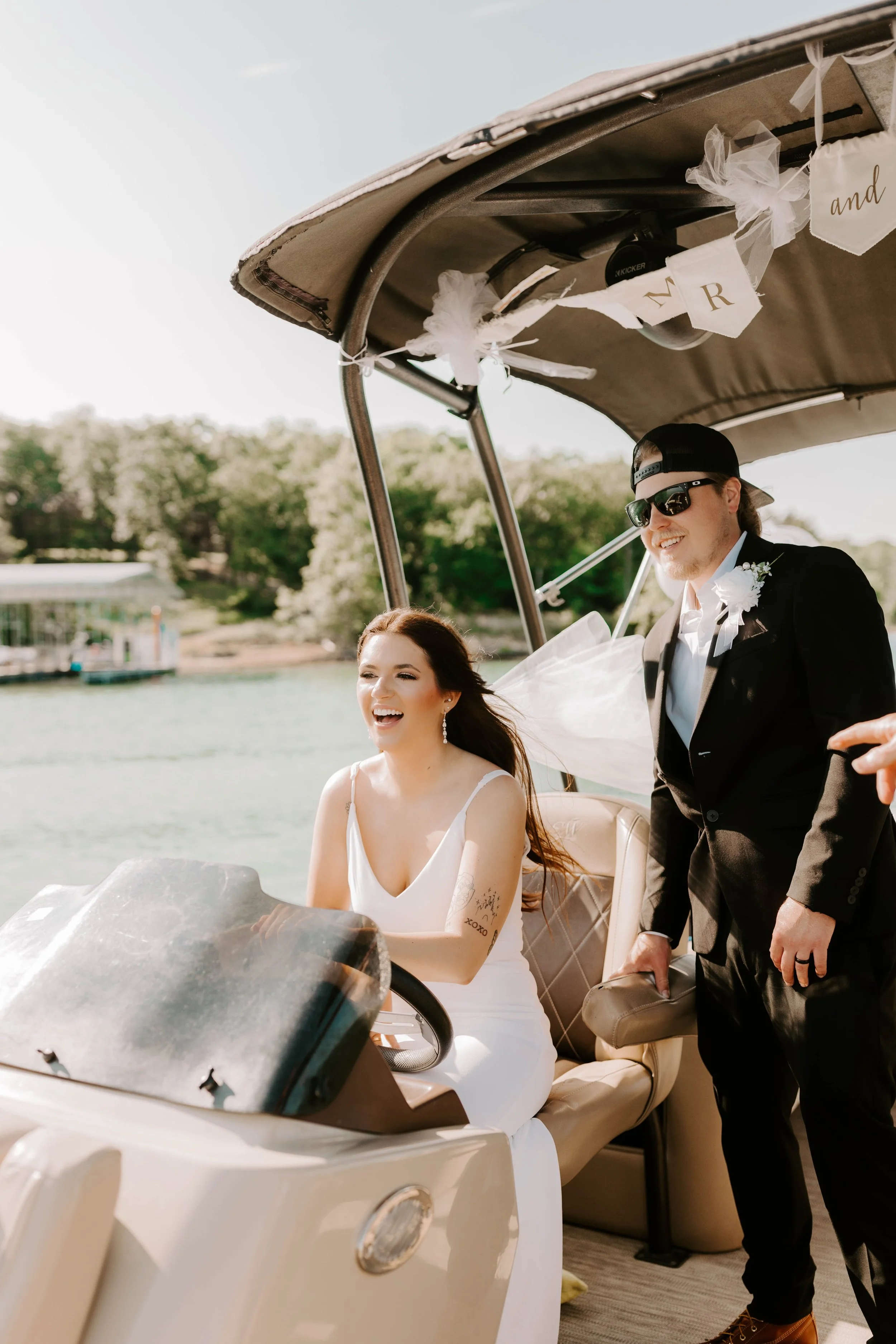 A woman in a white dress is sitting in a golf cart, smiling. A man in a black tuxedo, sunglasses, and a backward cap is standing beside her, also smiling. They are outdoors on a sunny day near a body of water with trees in the background.