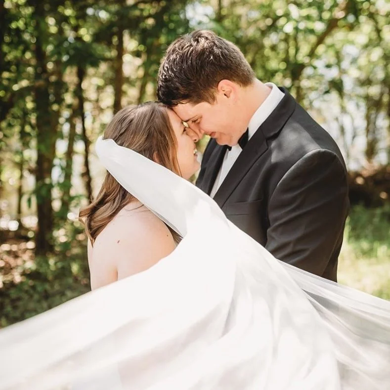 A bride and groom share a close moment outdoors, with the bride partially covered by her wedding dress and the groom in a black tuxedo leaning in.