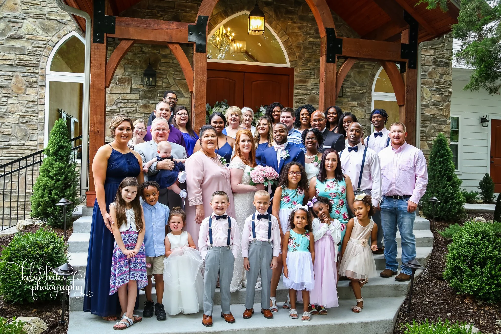 A large group of people, including adults and children, gathered on the steps of a stone and wood house for a family or wedding photo. The group is smiling and dressed in formal and semi-formal attire, with some children dressed in children's clothin